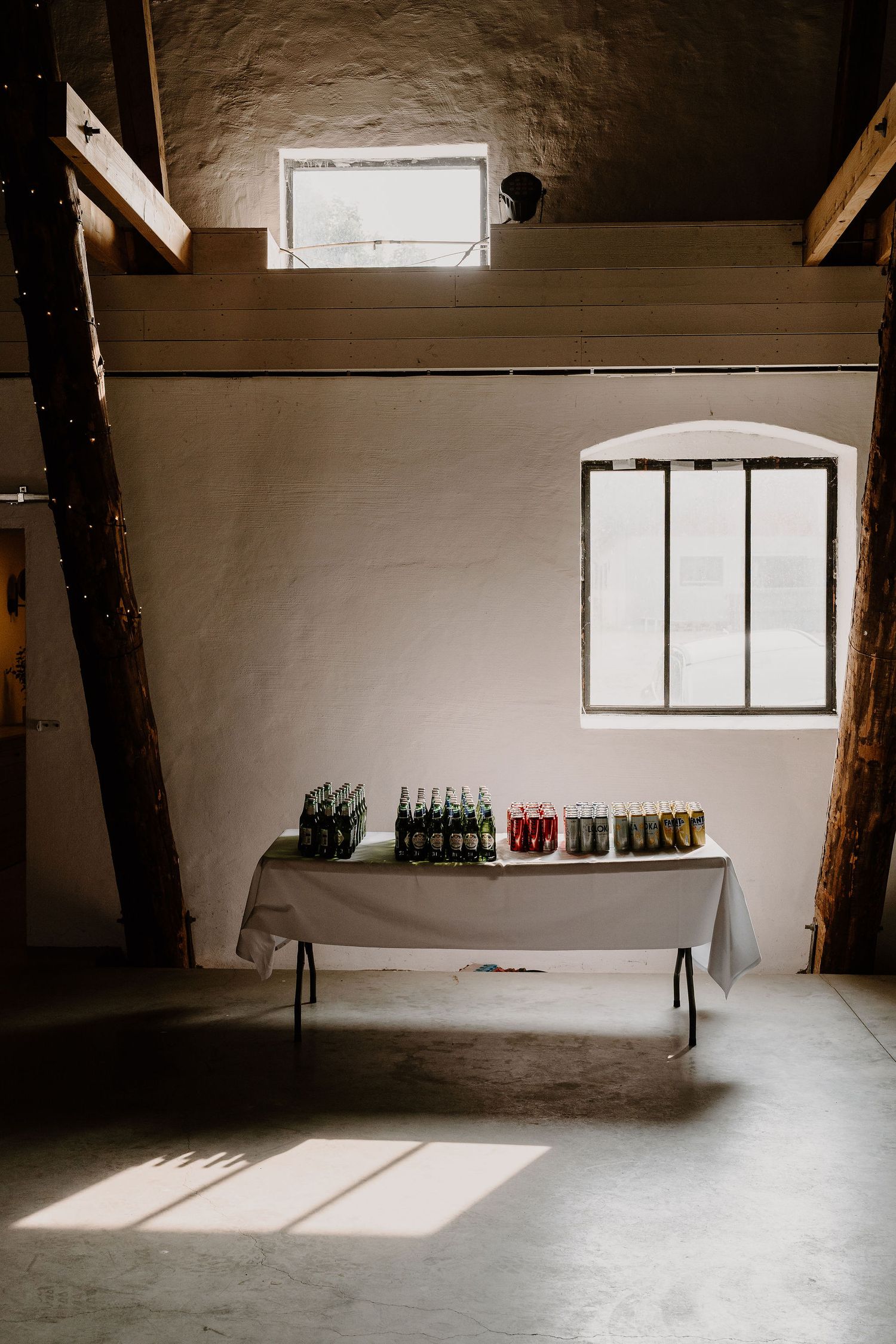 Minimalist industrial venue space with white walls, exposed beams, and bottles lined up on tables along concrete floors.