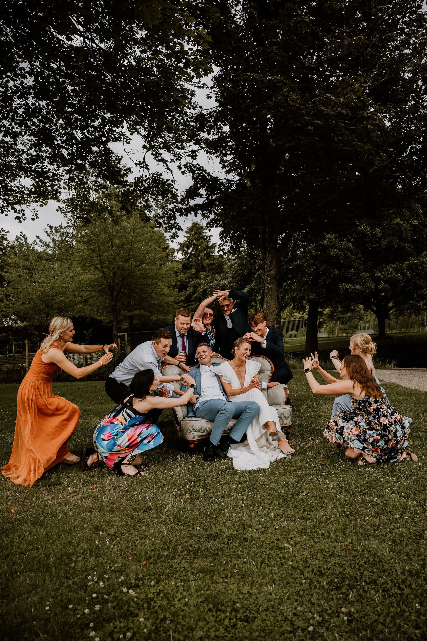 A playful group falls back in a domino effect while sitting on grass in a park setting on a summer day.