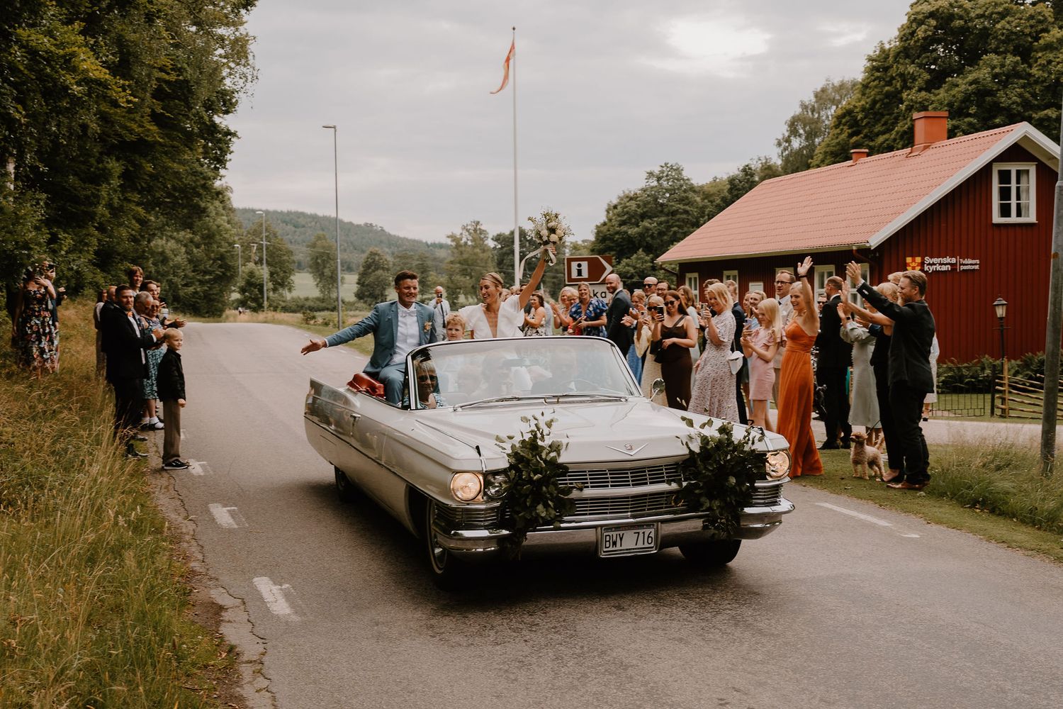 Classic white Cadillac convertible driving down a rural road with crowd cheering along both sides.
