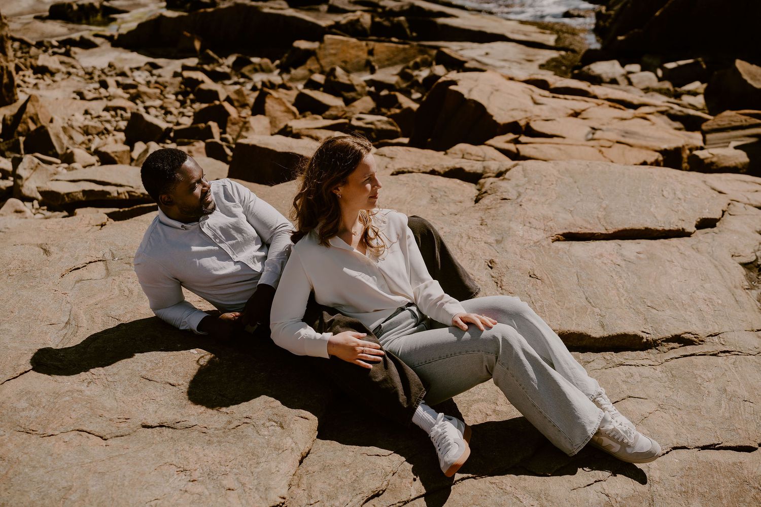 Two people in casual outfits relax together on sun-lit rocky terrain during an outdoor photo session.