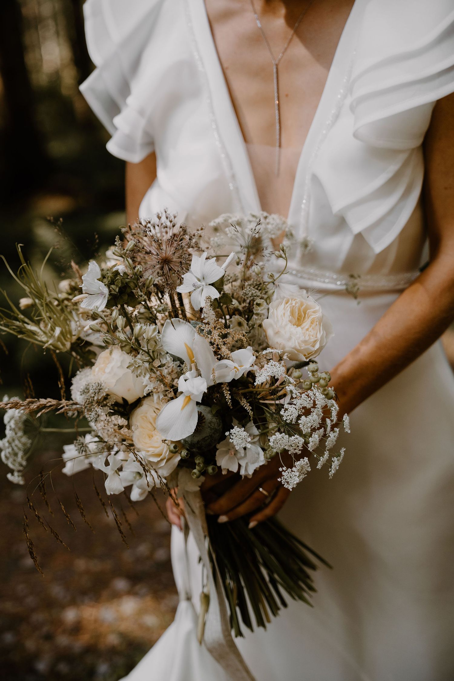 A delicate white bridal bouquet with roses and dried flowers held against a simple white wedding dress in natural light.