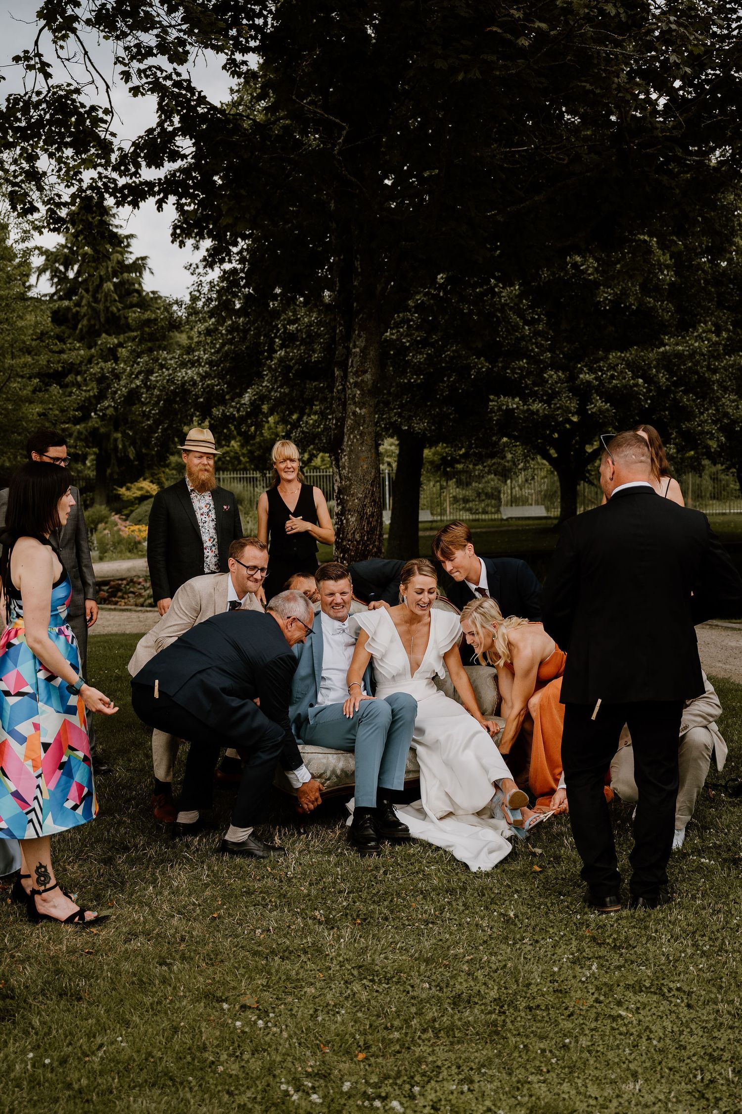 Wedding party sits casually on grassy hill having candid moments and laughing together outdoors.