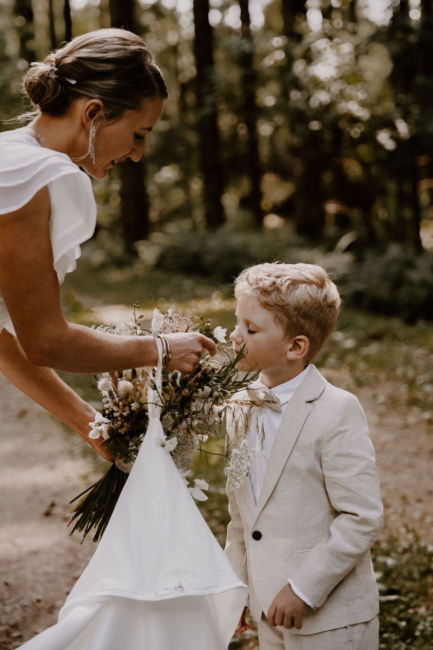 A touching moment between a person in a white wedding dress and a child in a light colored suit outdoors in a forest setting.