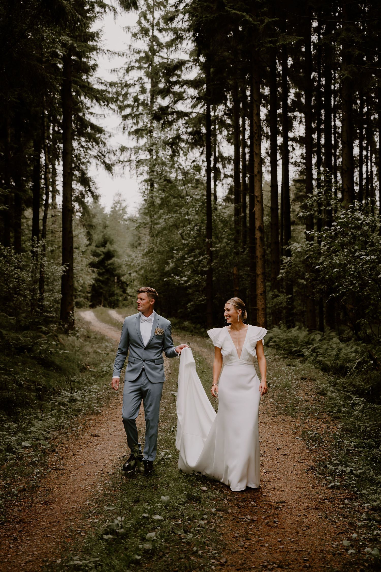 A couple in wedding attire walking together through a misty forest path surrounded by tall evergreen trees.