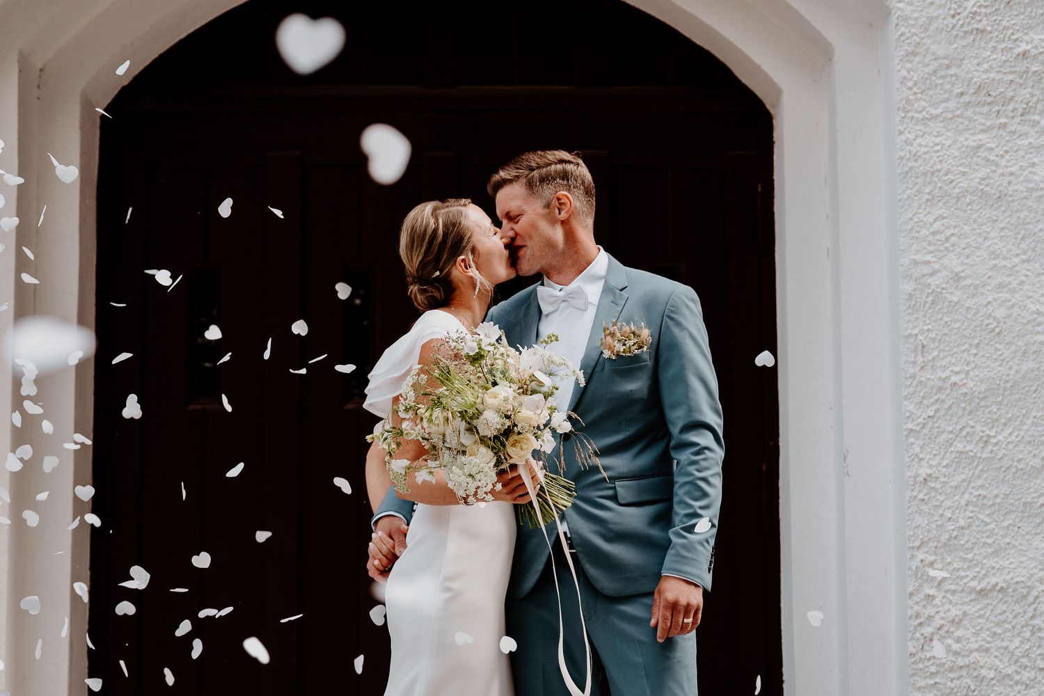 Newlyweds celebrate under shower of white confetti petals outside church archway.