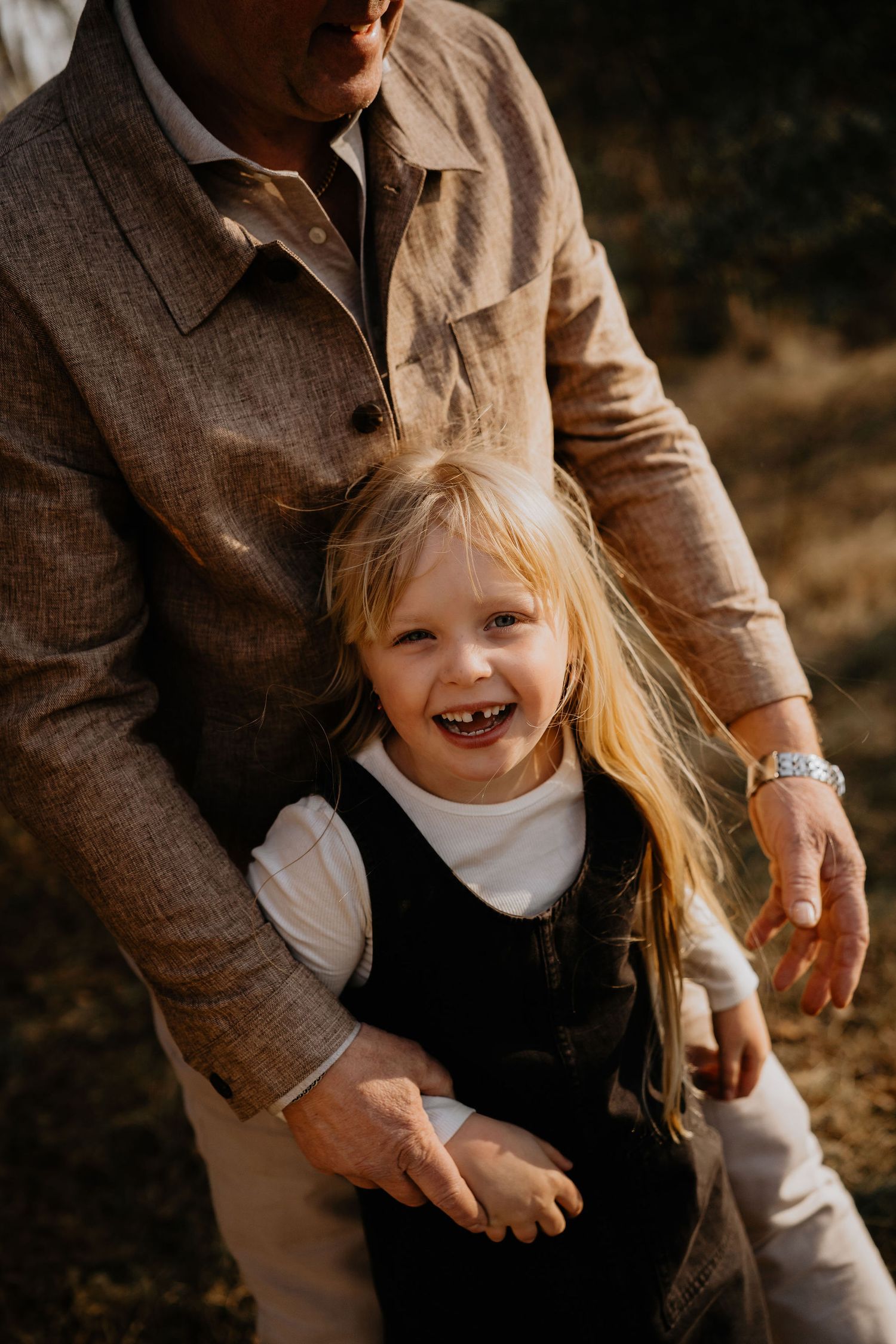 A joyful child with long blonde hair wearing a black dress laughs while being held by someone in a brown coat.