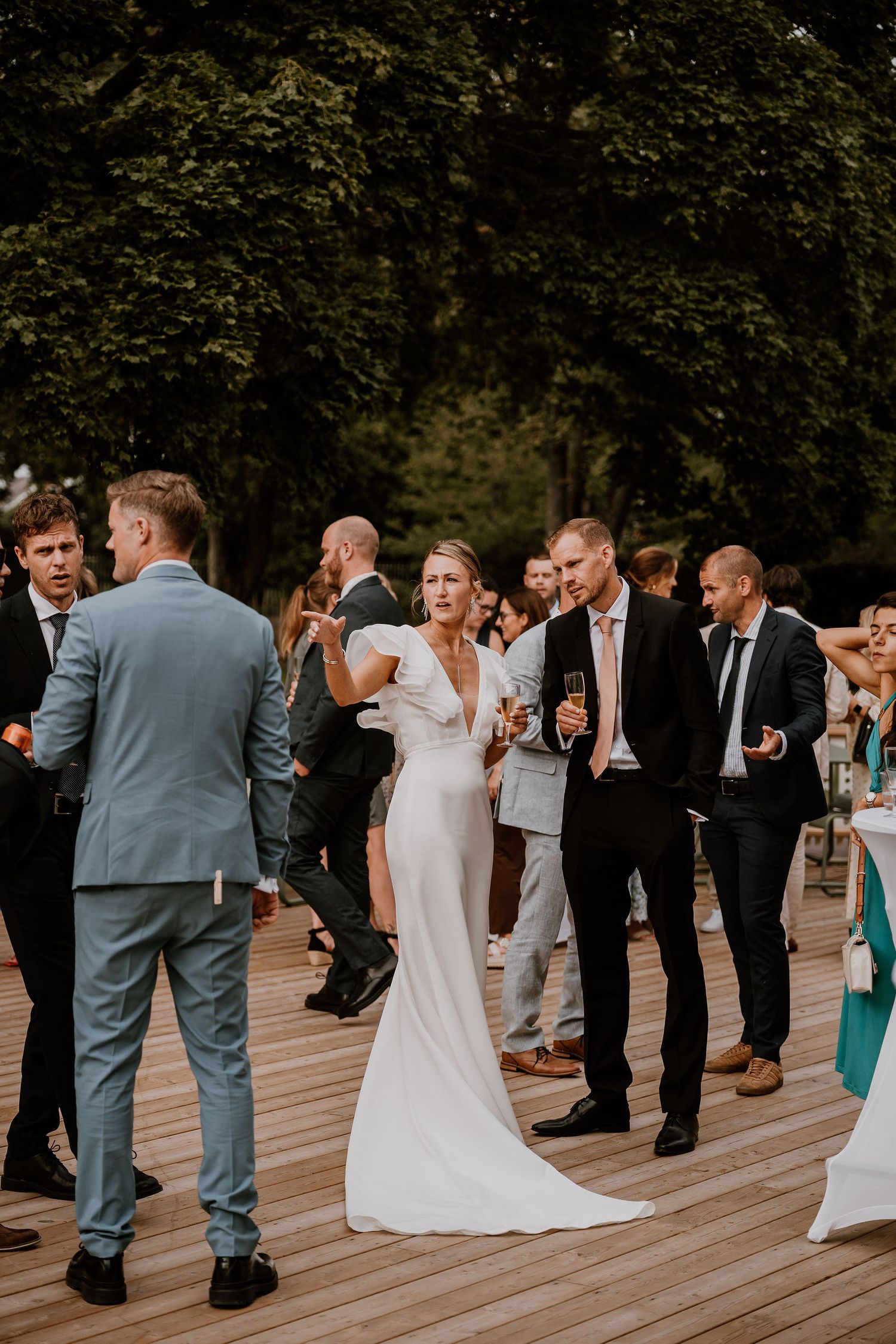 Wedding guests gathered on a wooden deck during an outdoor celebration with greenery in the background.