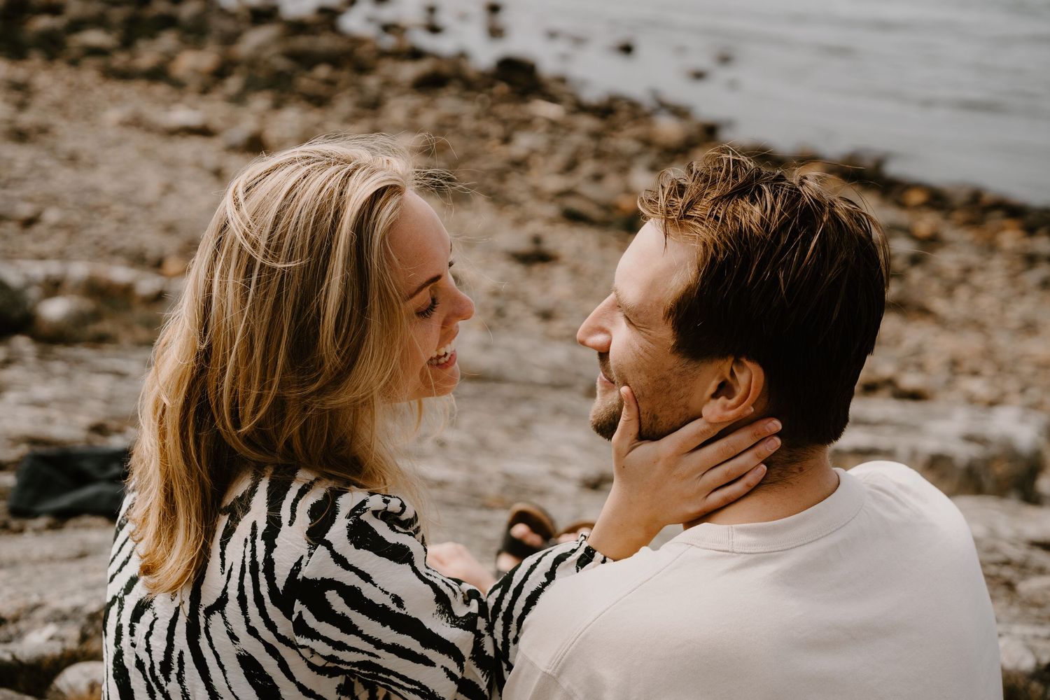 A couple shares an intimate moment together while sitting on a rocky beach at sunset.