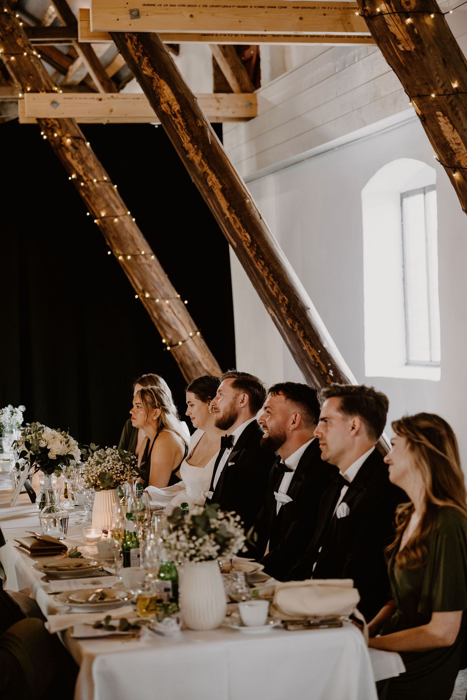 Wedding reception guests seated at a long white-clothed dining table under wooden beams with twinkle lights.
