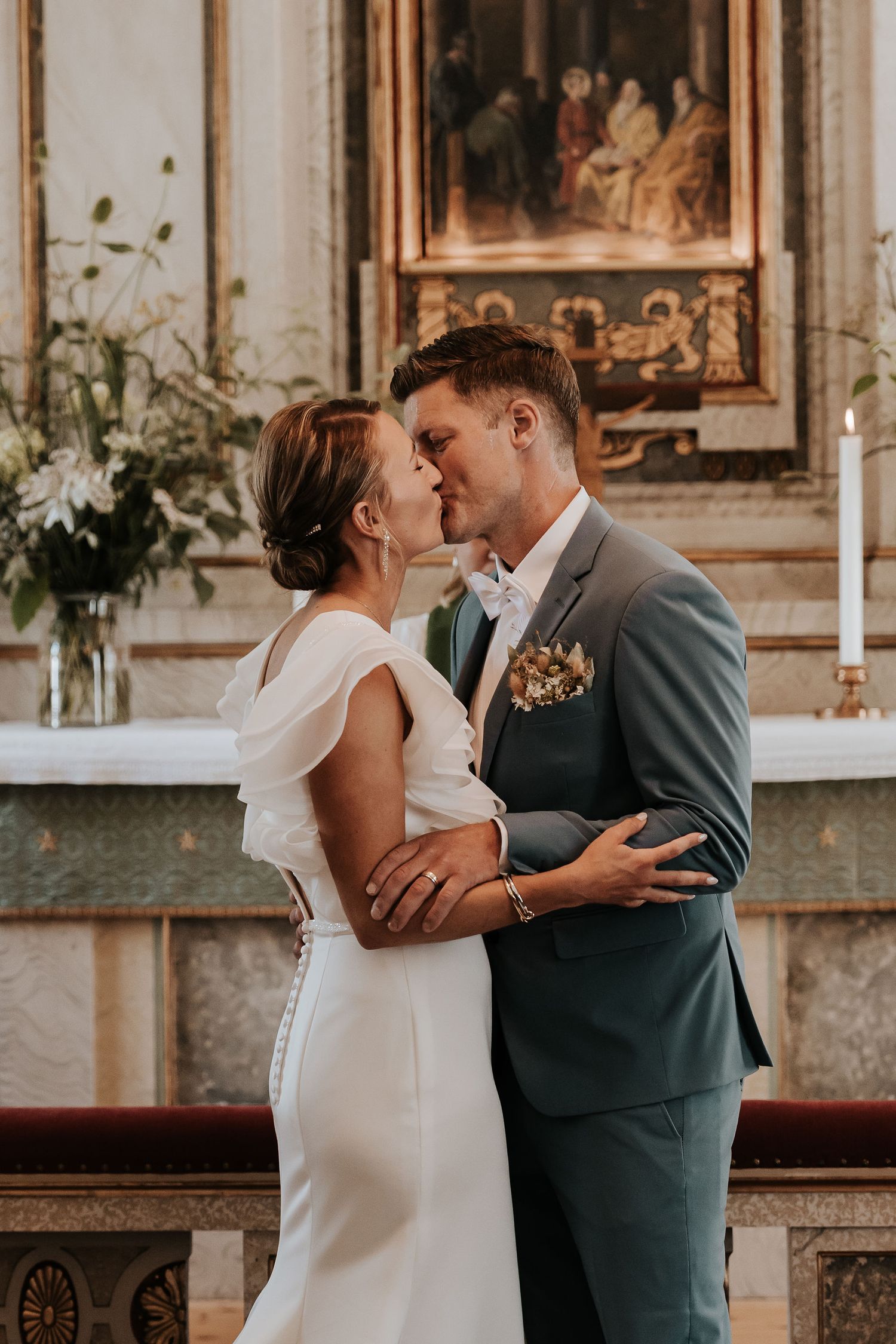 Romantic first kiss as newlyweds inside an ornate church sanctuary with gold frames and white floral arrangements.