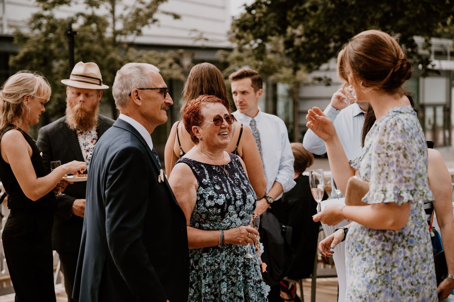 Guests at a formal outdoor event mingle and socialize while standing in small groups on a sunny day.