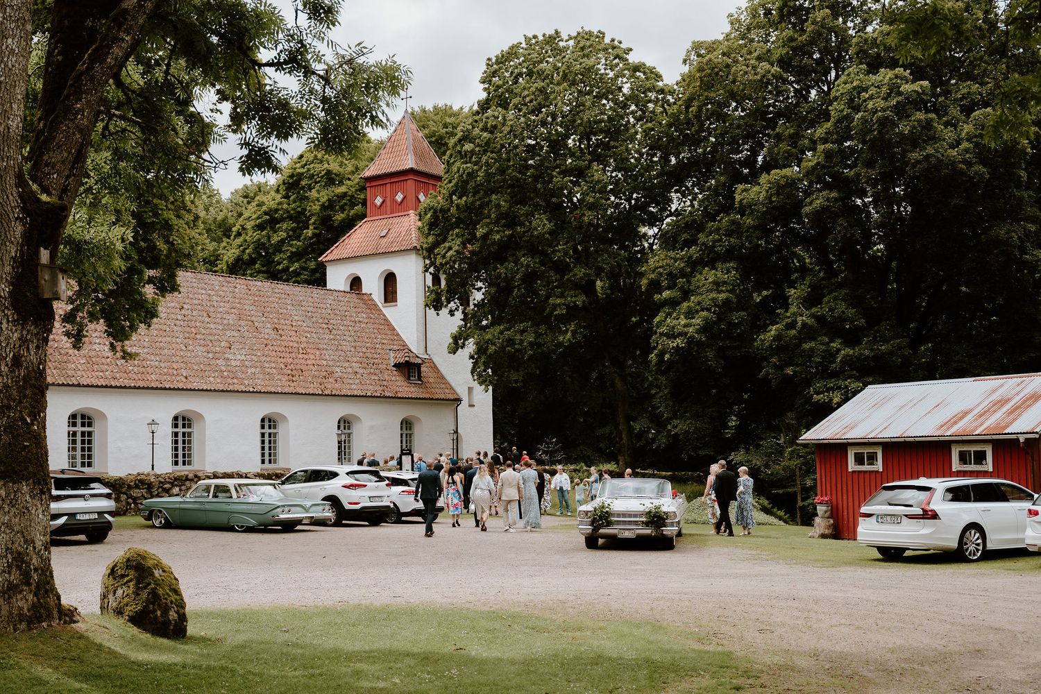 Classic cars parked outside a white church with bell tower and red barn on a sunny summer day.