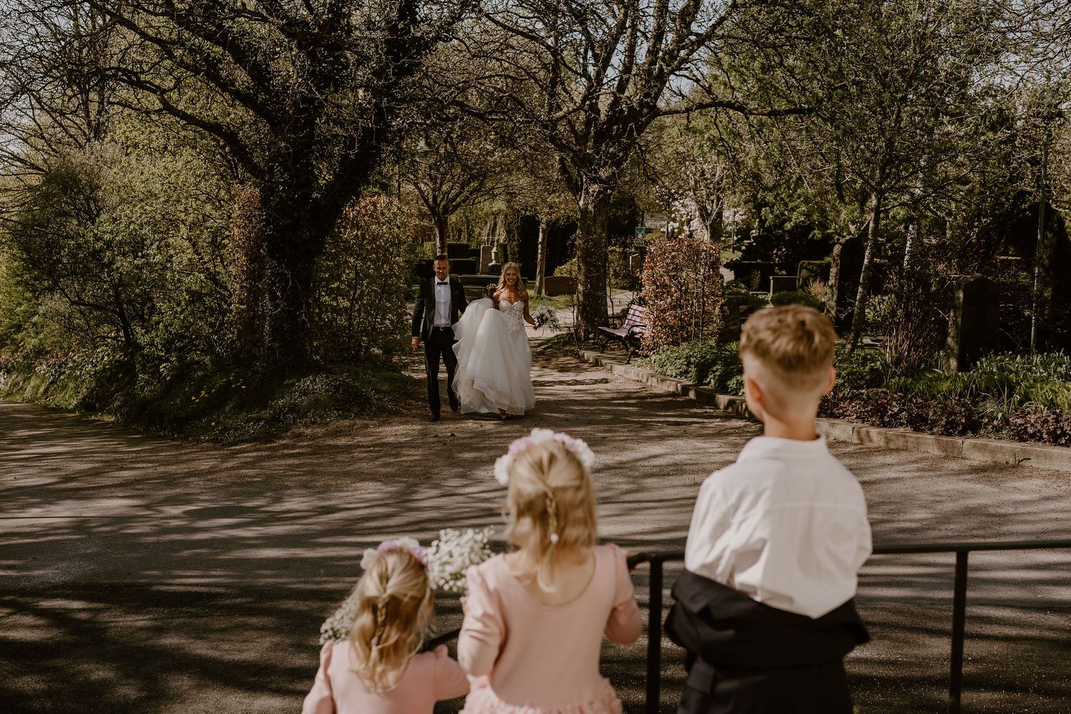 Three children watch figures walking down a sunlit garden path lined with trees on a warm spring day.