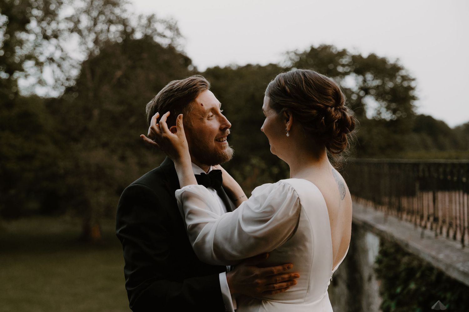 A tender romantic moment between a couple in wedding attire standing in front of a rustic bridge at dusk.