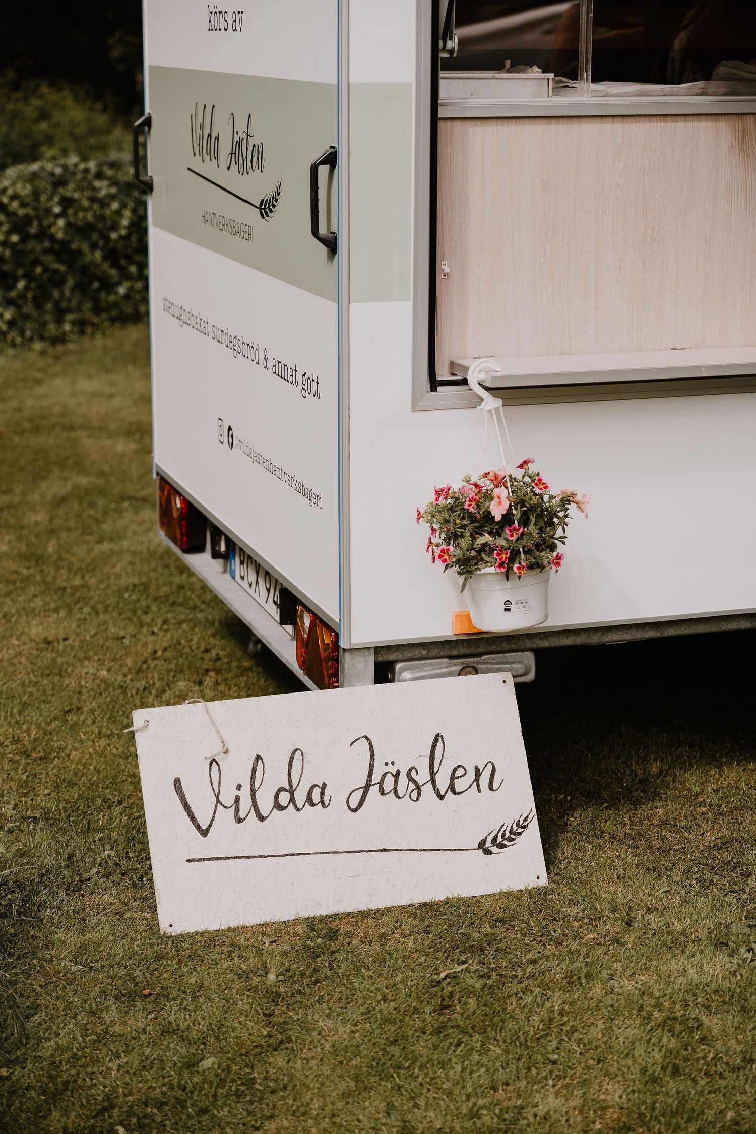 White food truck with pink flowers and a handwritten 'Vilda Fasken' sign parked on grass.