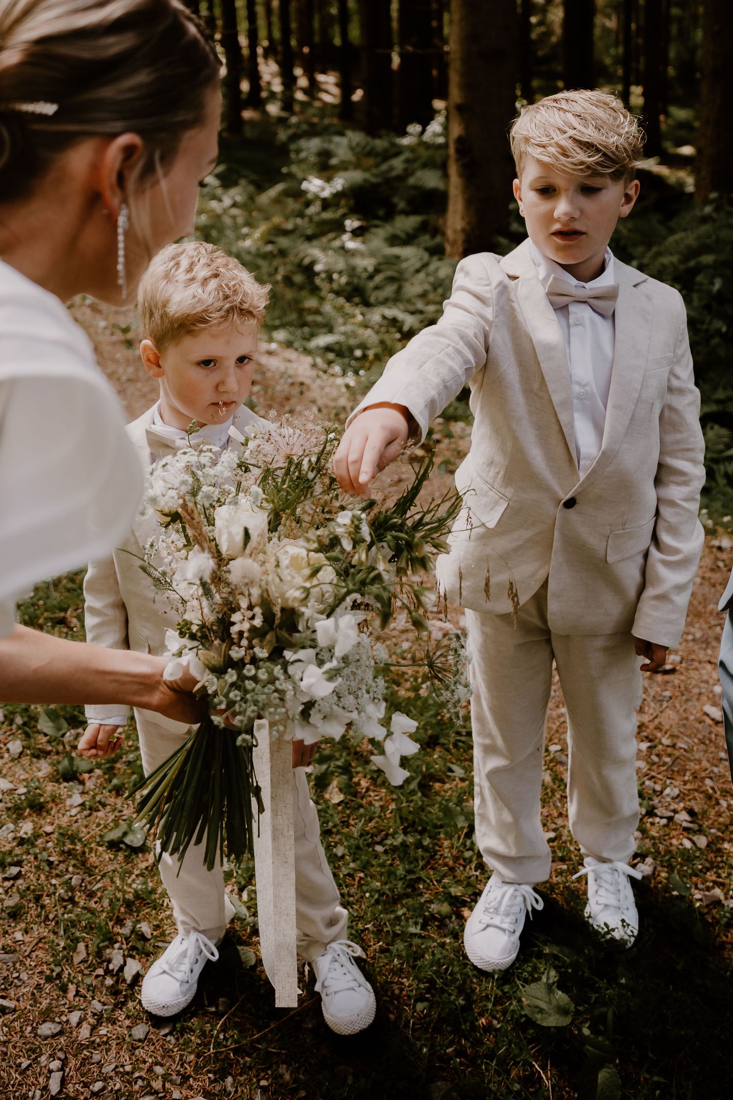 Small ring bearer in light grey suit and bow tie poses with flowers in outdoor woodland setting.