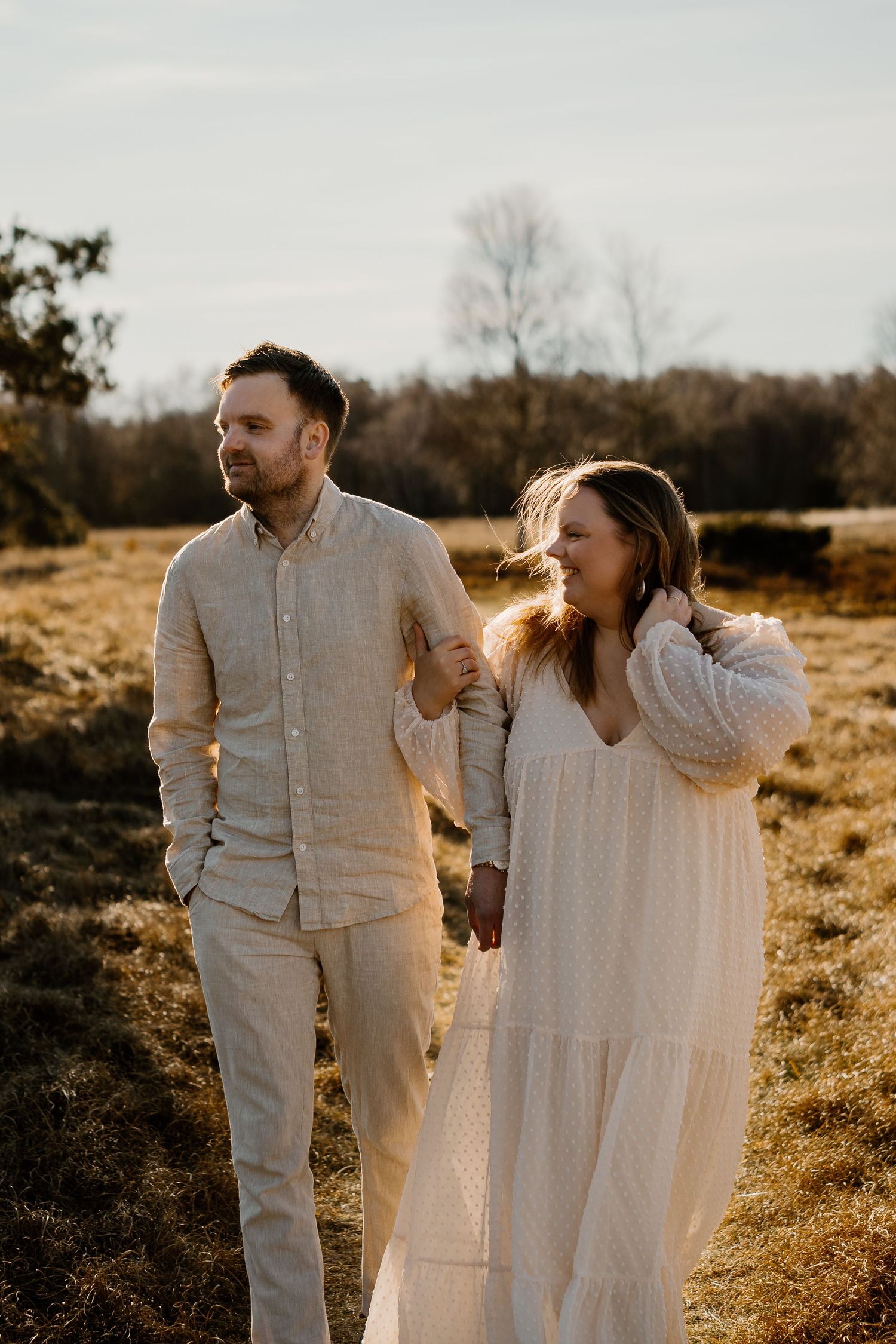 A couple in white outfits holding hands walks through a sunlit field at golden hour during a romantic photoshoot.