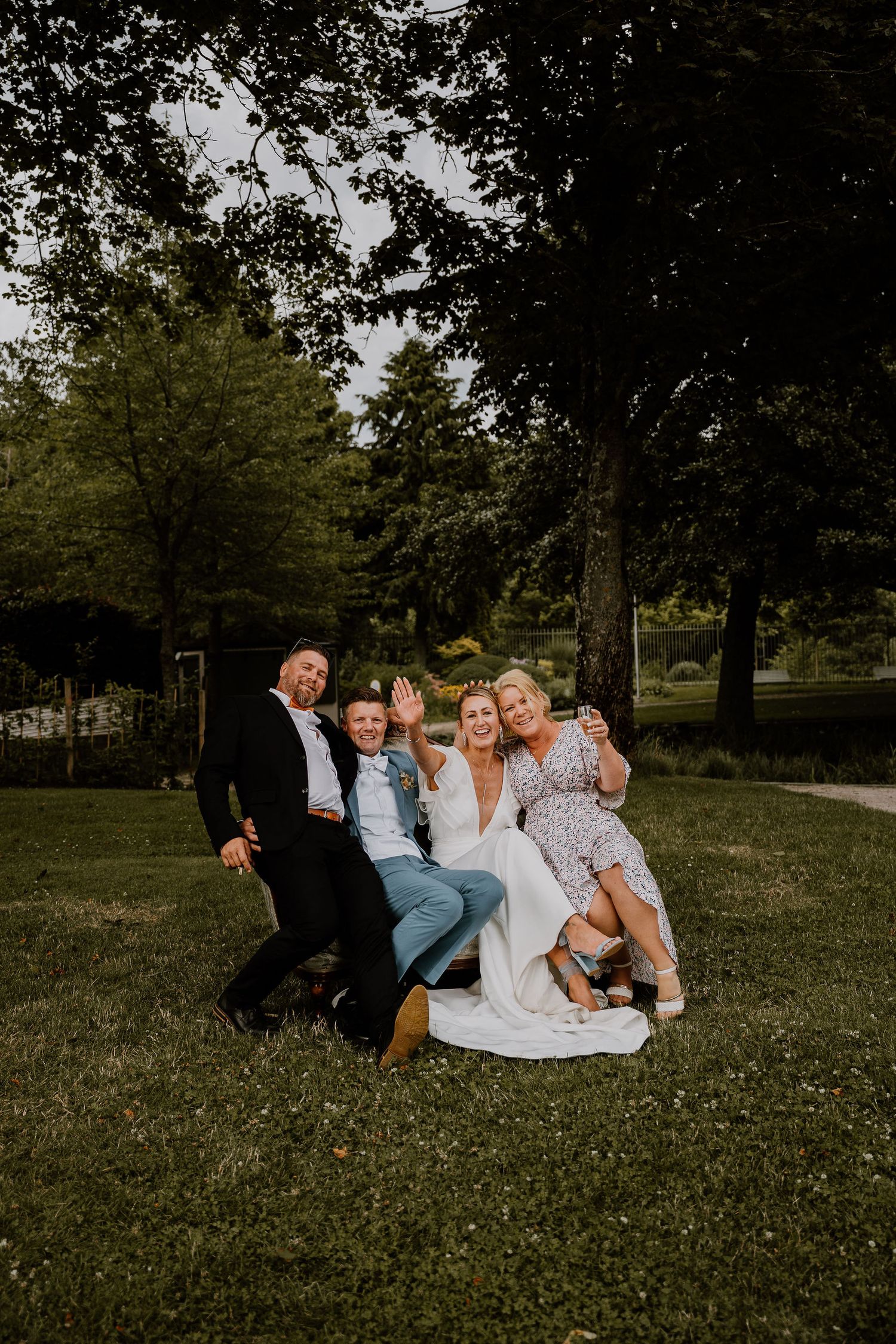 A group relaxes together on a grassy lawn under trees in a casual outdoor photoshoot setting.