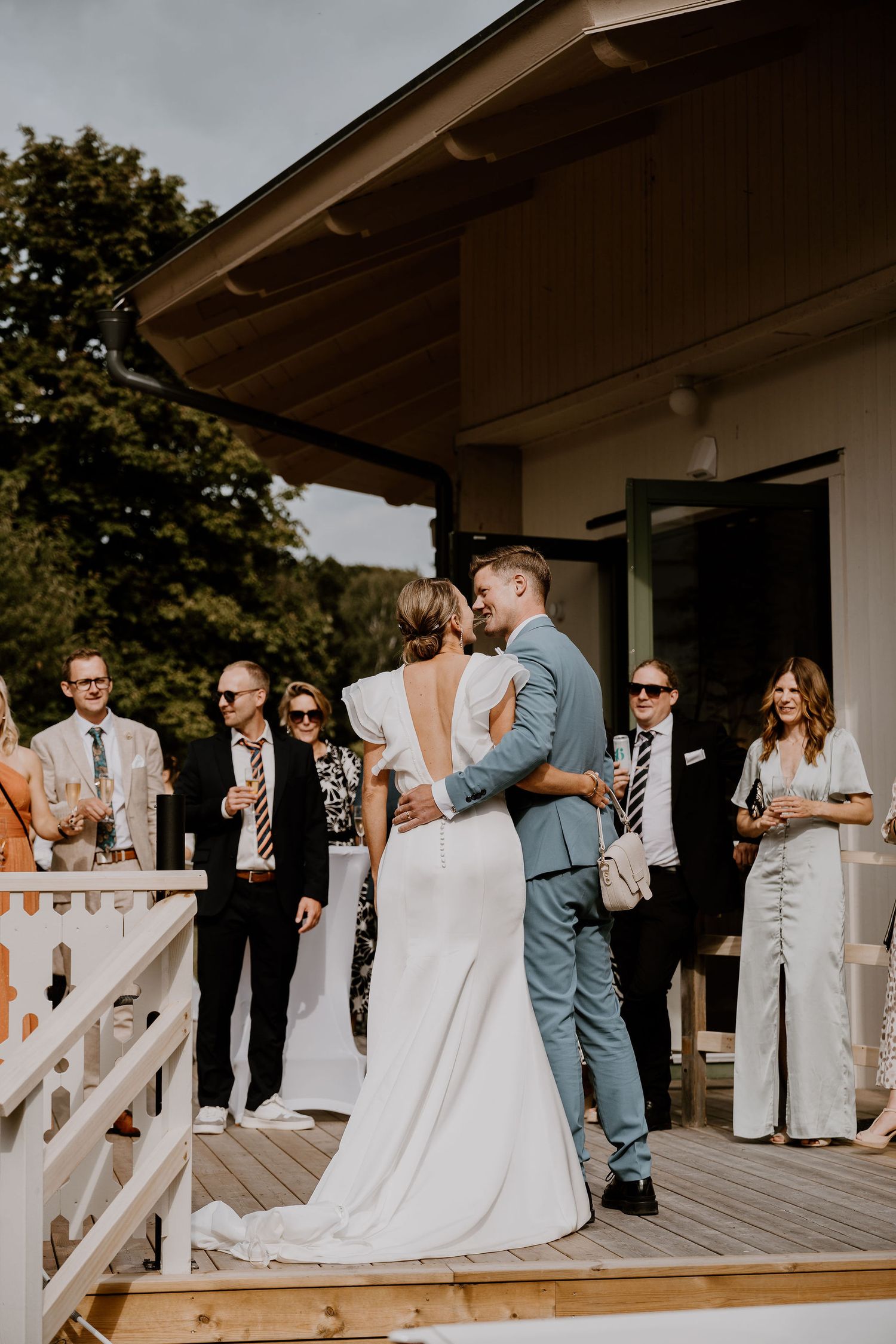 Wedding guests gather on a wooden deck to celebrate as the newlyweds share a romantic kiss during the outdoor ceremony.