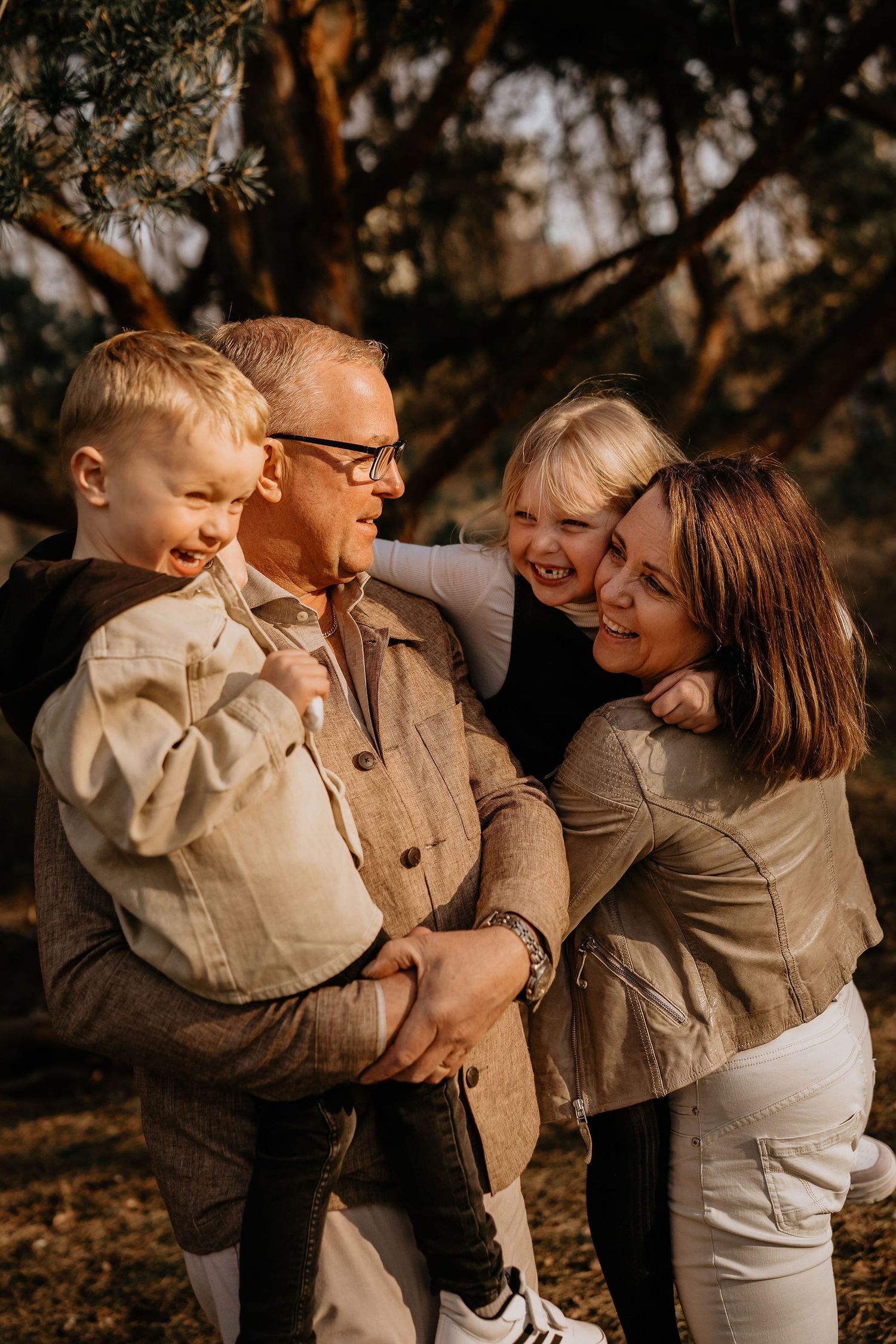 A candid outdoor family portrait shows four people embracing and laughing together in warm autumn lighting.
