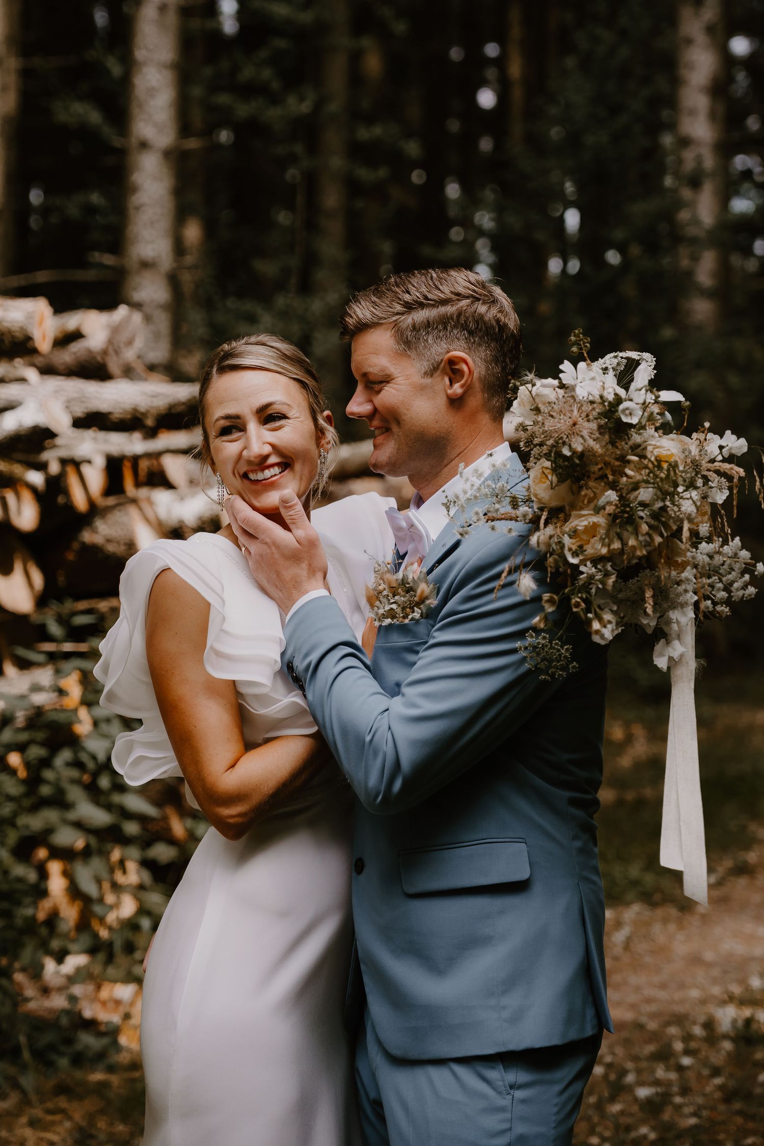 A happy couple laughs together during a forest wedding ceremony while holding white floral bouquets.