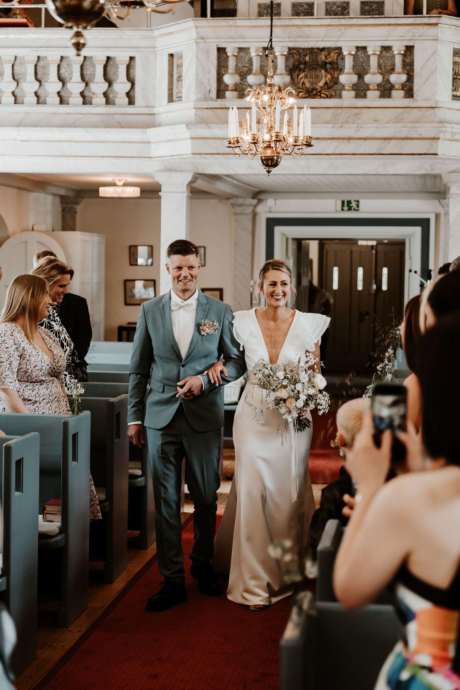 A wedding ceremony processional in an elegant church with white walls and chandeliers.