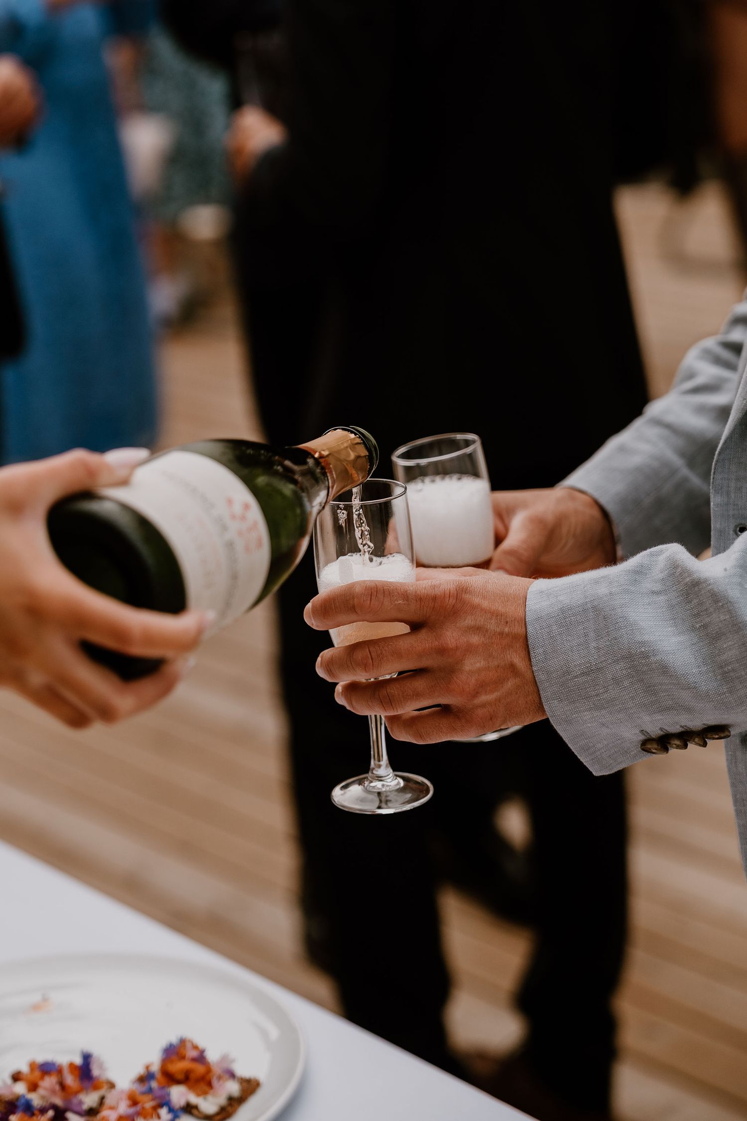 People holding champagne glasses and wine during a social gathering with wooden table visible in the background.
