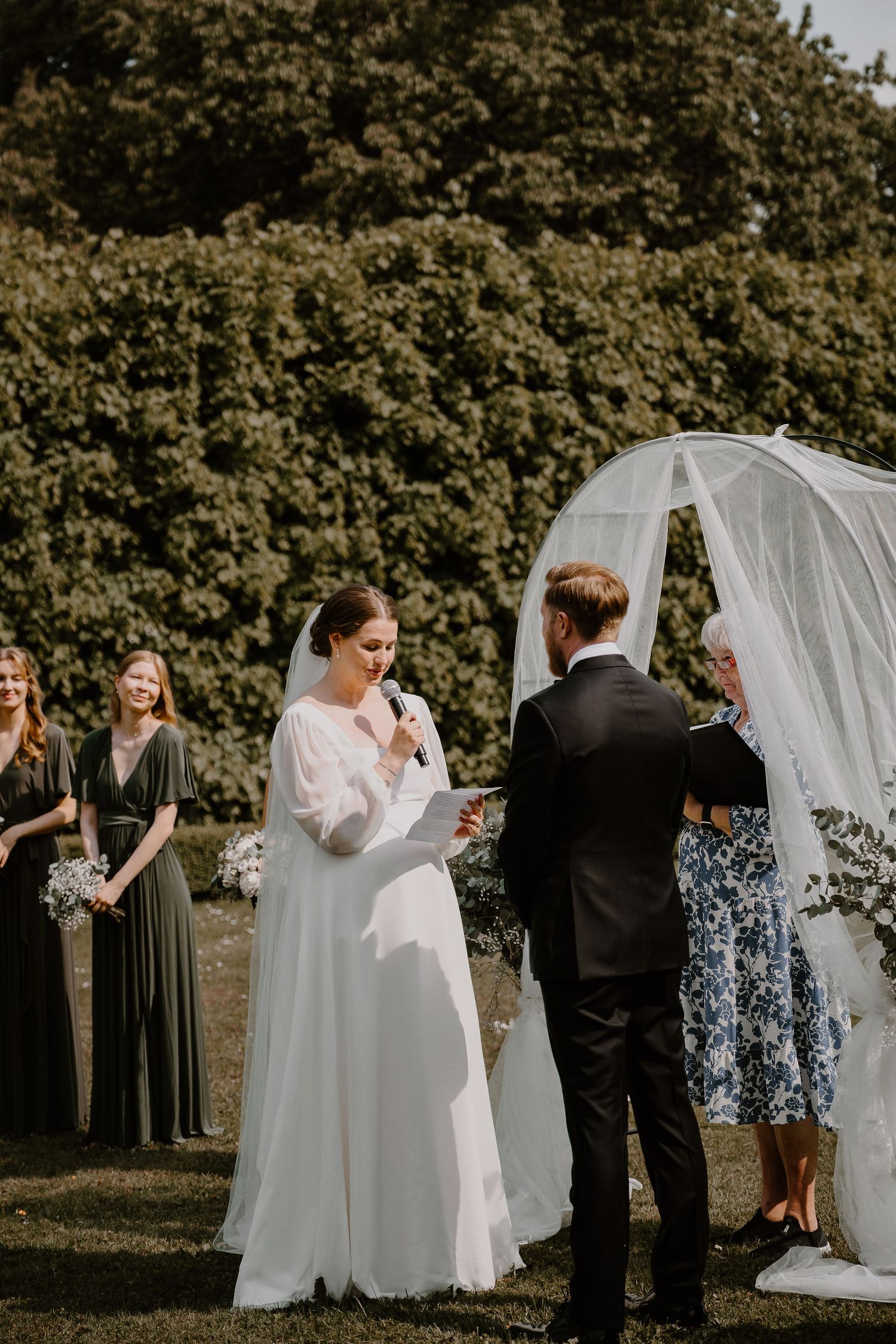 An outdoor wedding ceremony takes place under a white arch with bridesmaids in green dresses and floral decor.