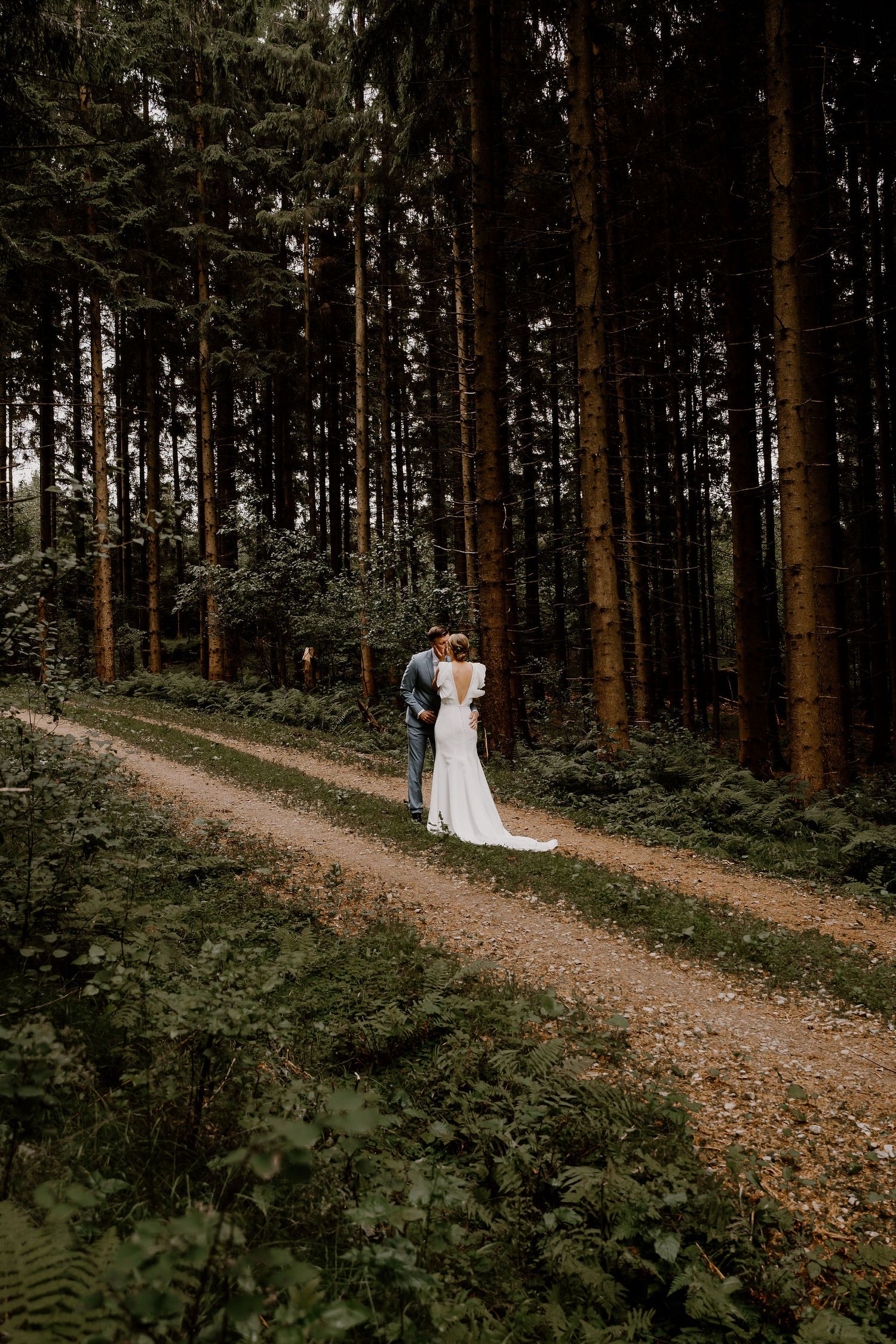 Wedding couple walks hand in hand along a winding forest path surrounded by tall pine trees and ferns.