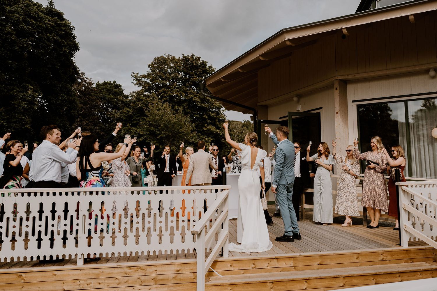 Wedding guests gather outside a residential venue with white picket fencing during a celebration.