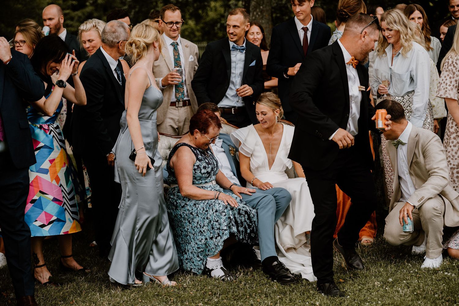 Large group of wedding guests sit casually on grass during outdoor summer celebration.