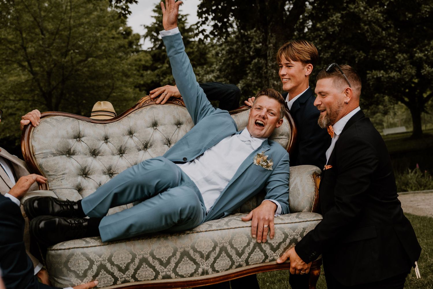 Wedding party lounges casually on vintage tufted sofa during outdoor garden photoshoot.