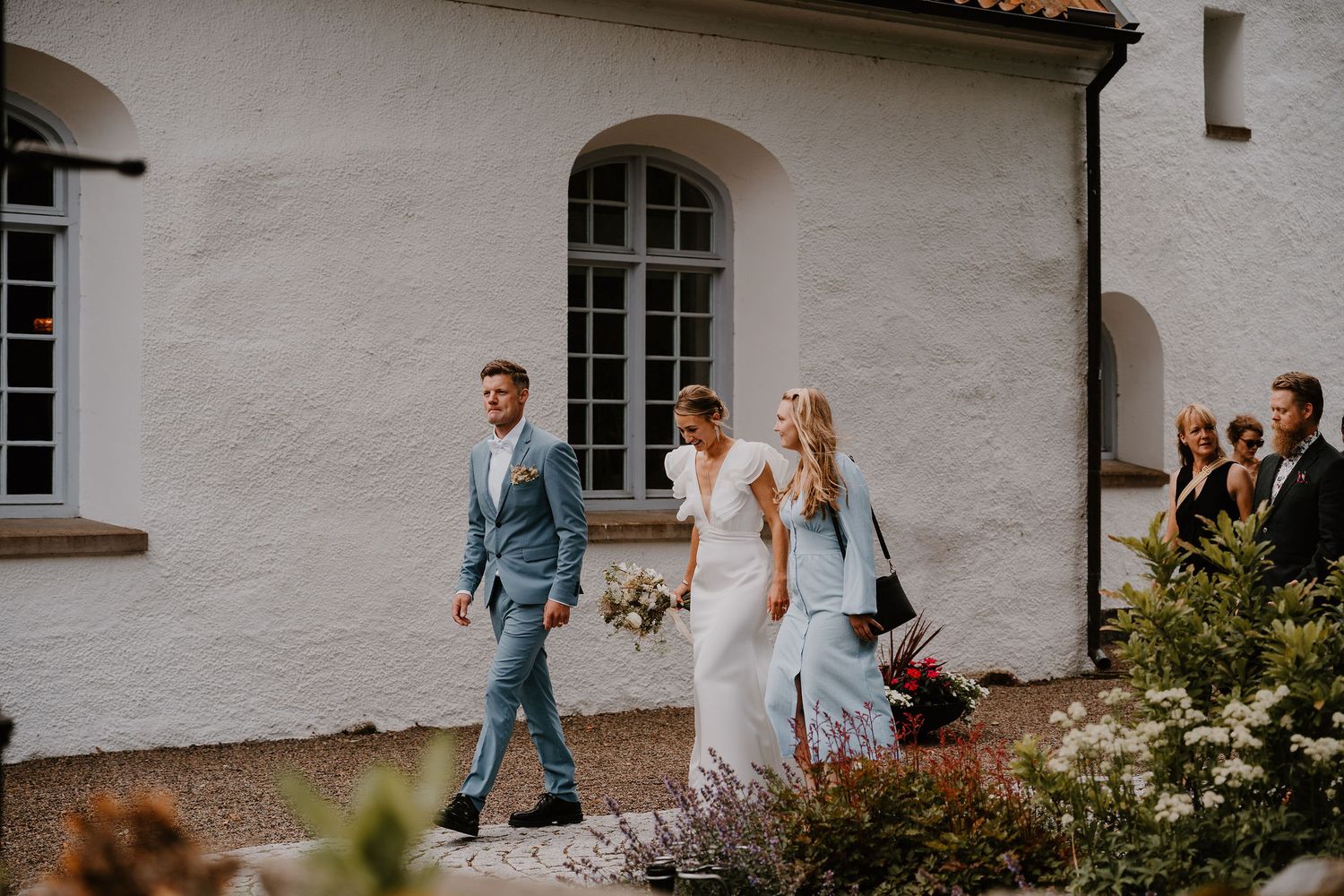 Wedding party walks along pathway by white stucco building with arched windows on sunny afternoon.