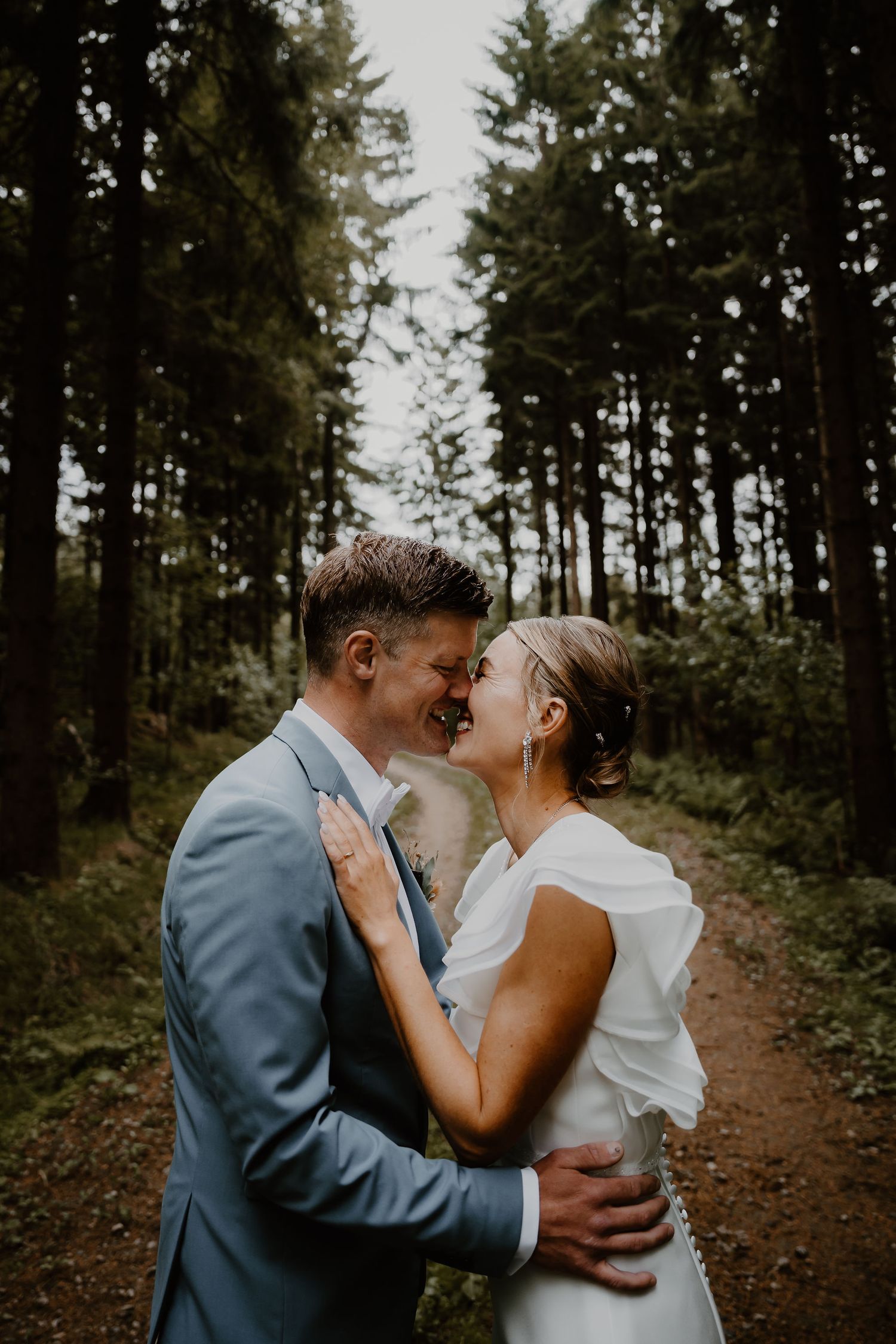 A romantic wedding couple shares an intimate moment on a wooded forest path, captured in moody natural lighting.