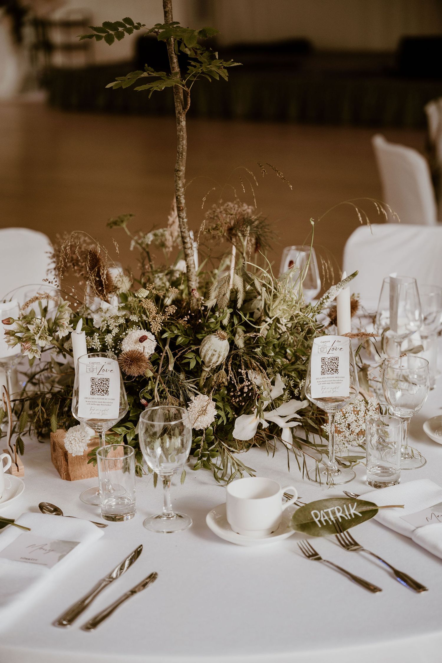 Elegant wedding reception table setting with white linens, wine glasses, and natural greenery centerpieces.
