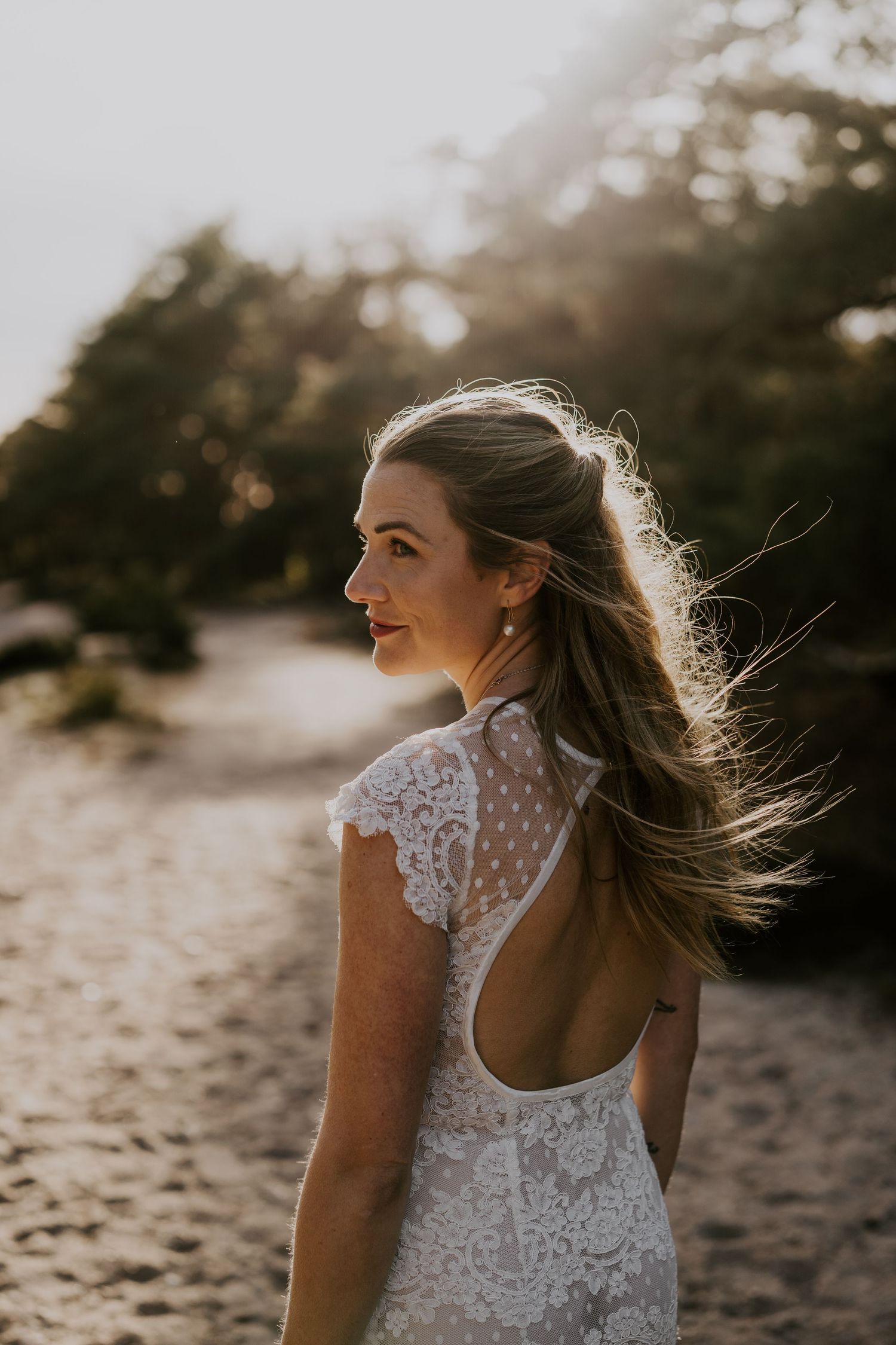 A bride in a backless lace wedding dress stands on a beach at sunset with sunlit hair blowing in the wind.