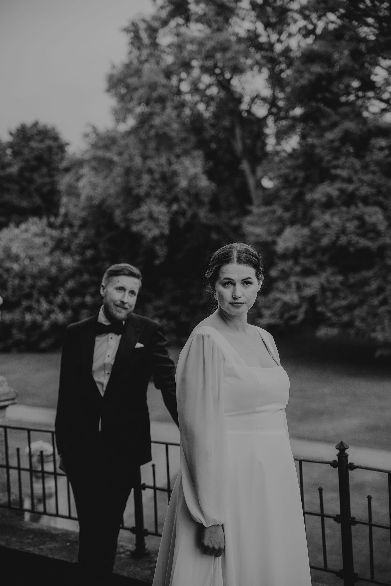 Black and white wedding portrait of a couple standing by an iron railing, surrounded by trees in a garden setting.