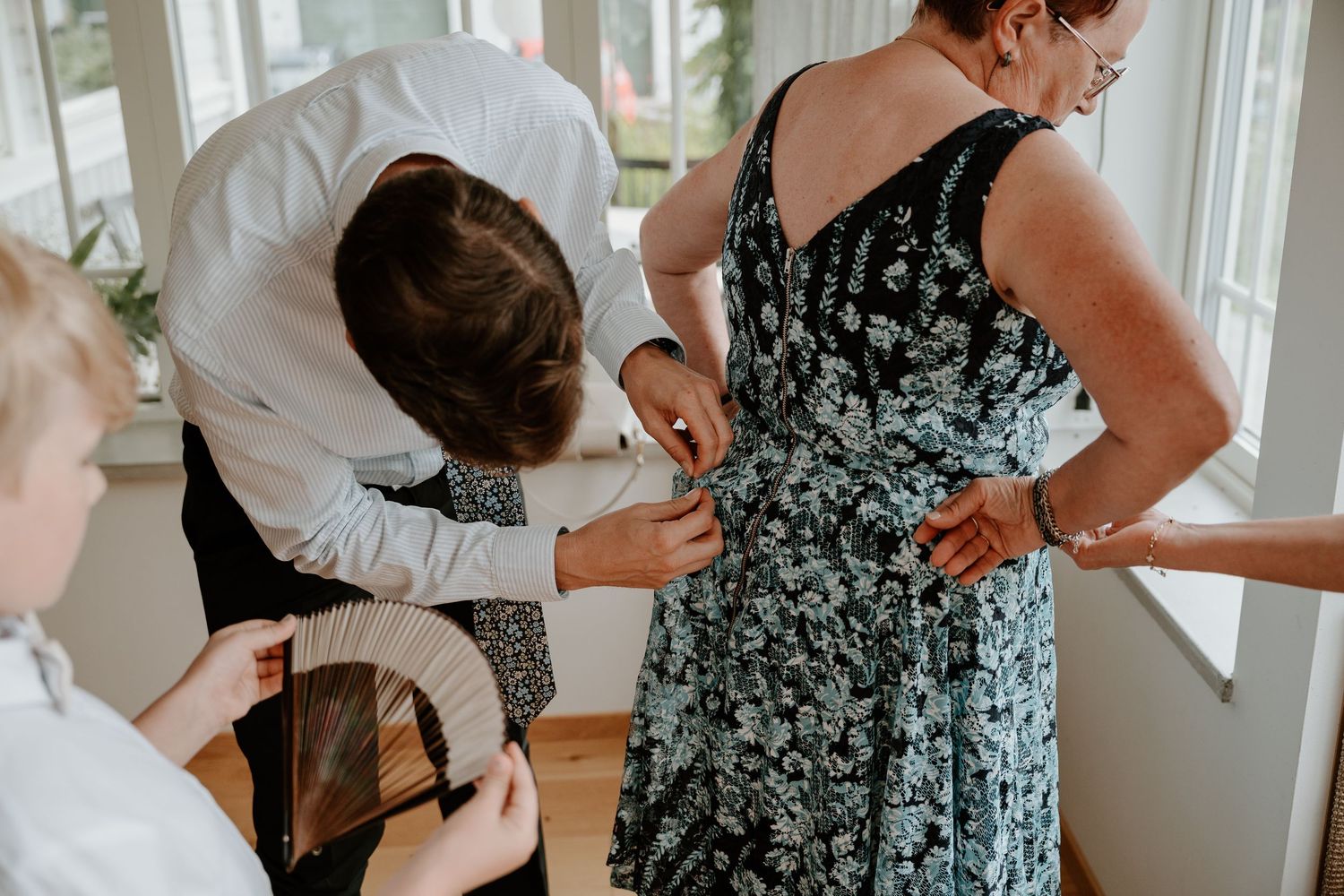 People getting dressed and helping each other prepare for a formal event in a bright, sunlit room.