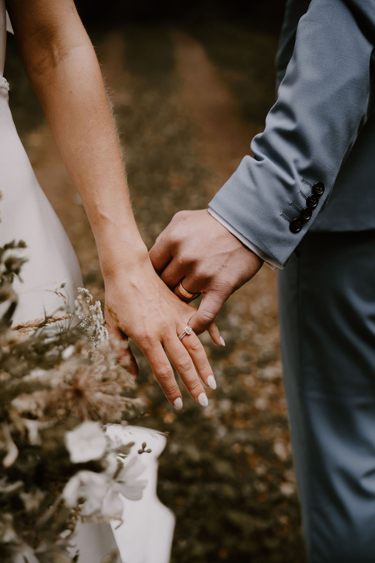 Close-up shot of newlywed couple holding hands against dark natural background showing wedding attire details.