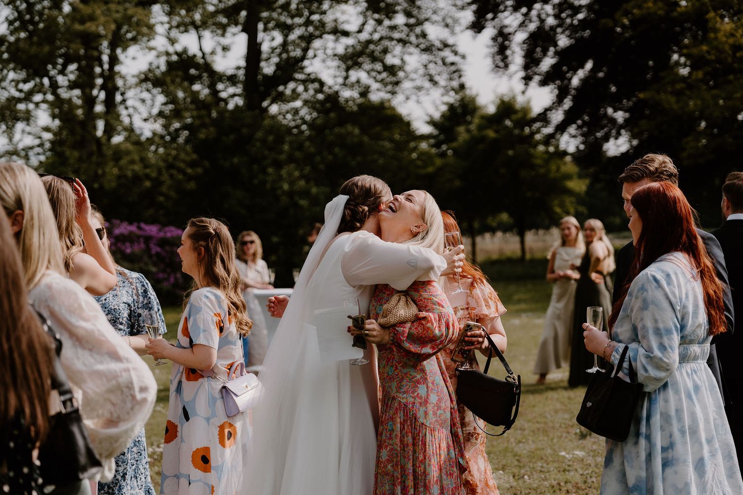Wedding guests gather on a sunny lawn to celebrate as the couple embraces during an outdoor ceremony reception.