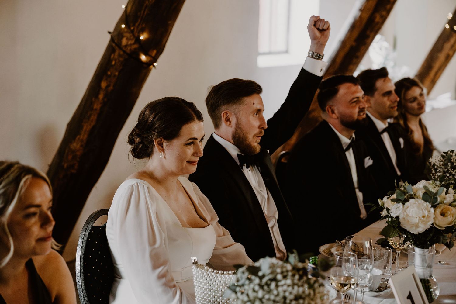 Wedding party seated at reception table raise their hands in celebration at rustic venue with exposed wood beams.