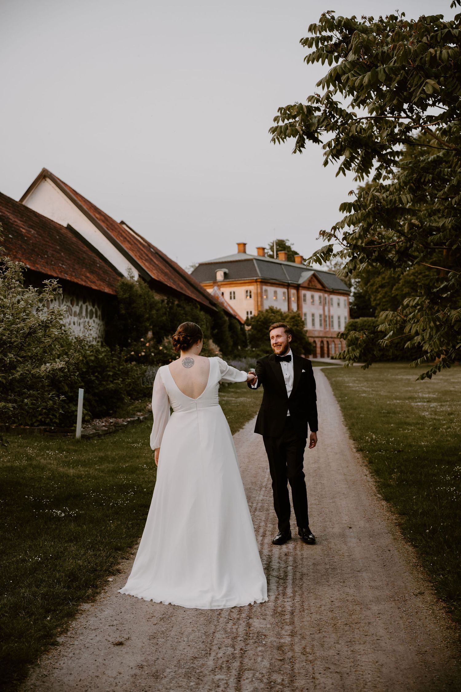 A couple walking down a dirt path through green grounds in timeless black and white wedding attire.
