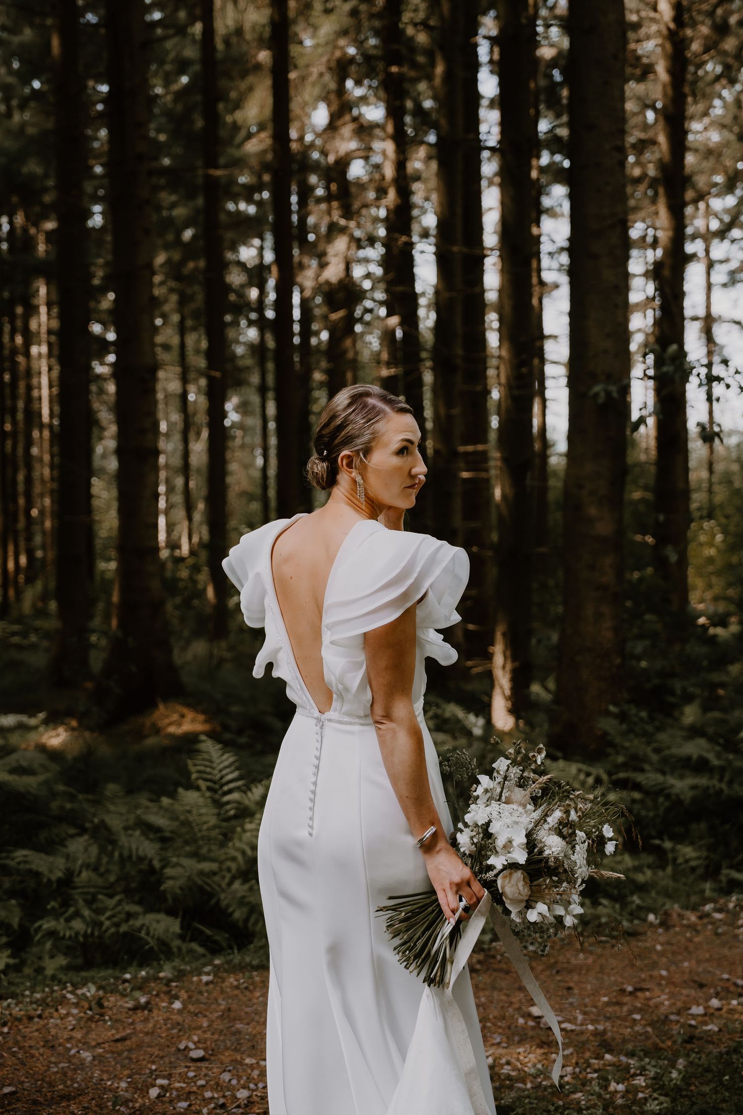A bride in a flowing white dress stands among tall pine trees in a moody forest setting.