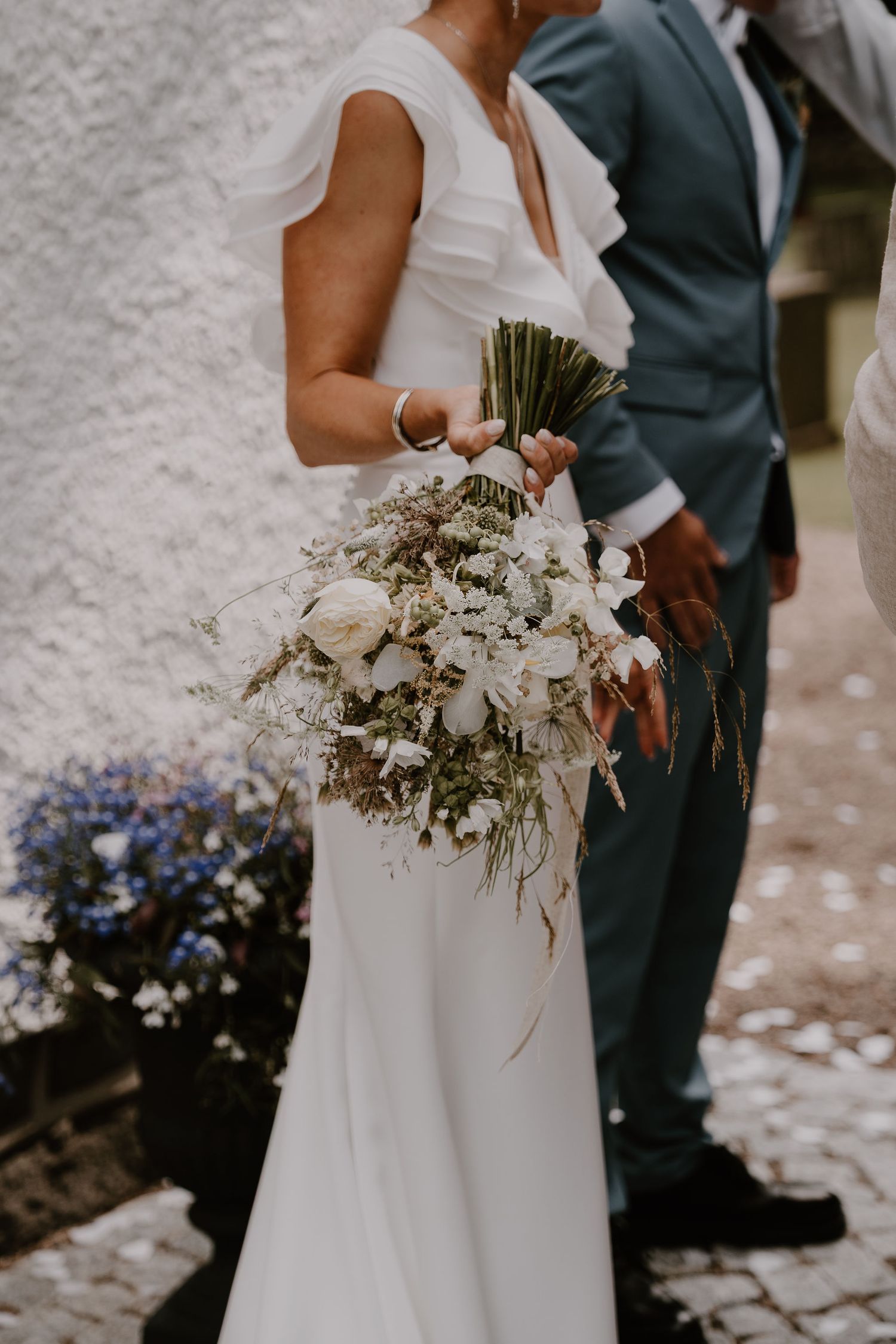 White wedding bouquet with delicate flowers and greenery held against a flowing white dress at an outdoor ceremony.