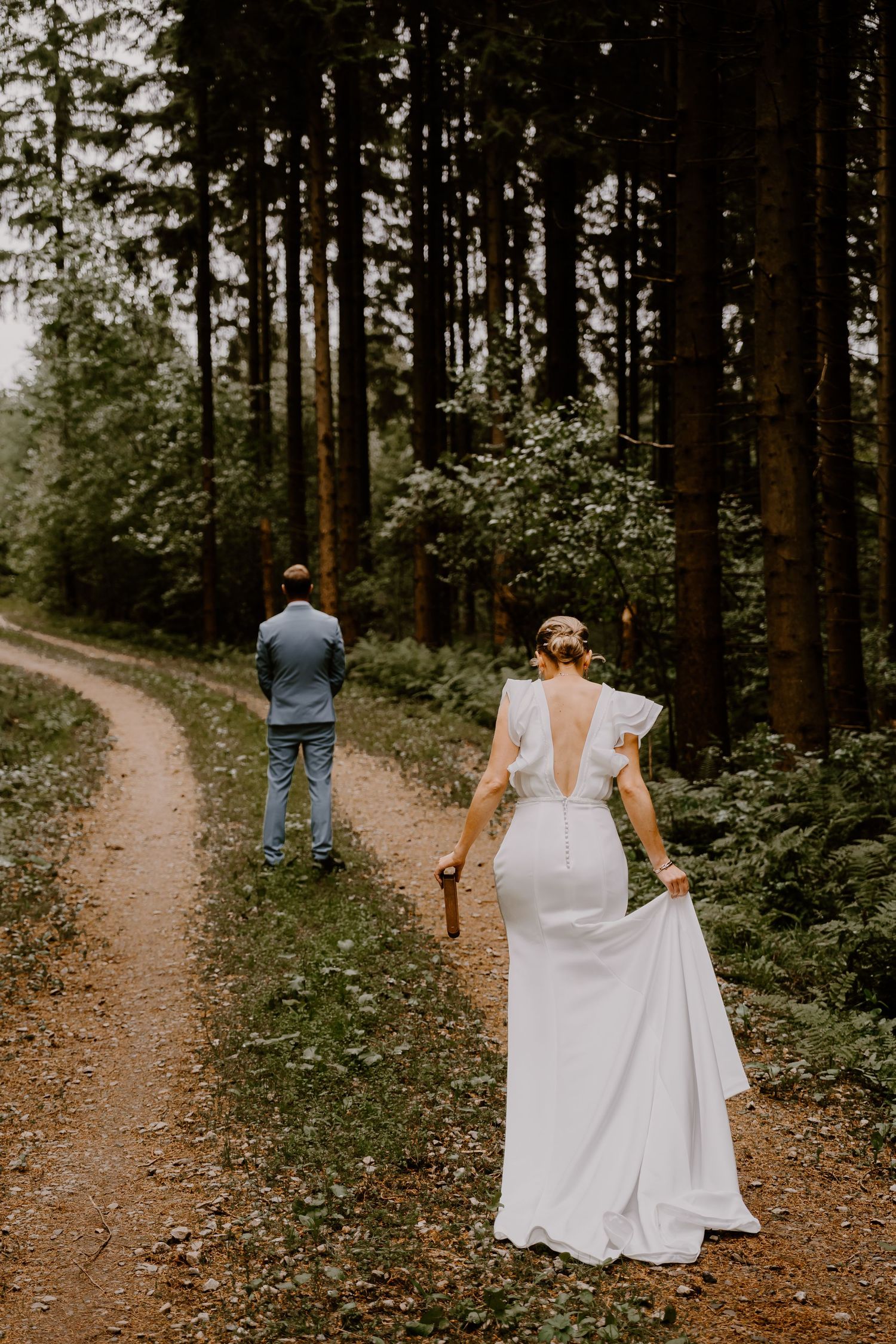 Two people stand on a winding forest path surrounded by tall evergreen trees, with one wearing a white wedding dress.