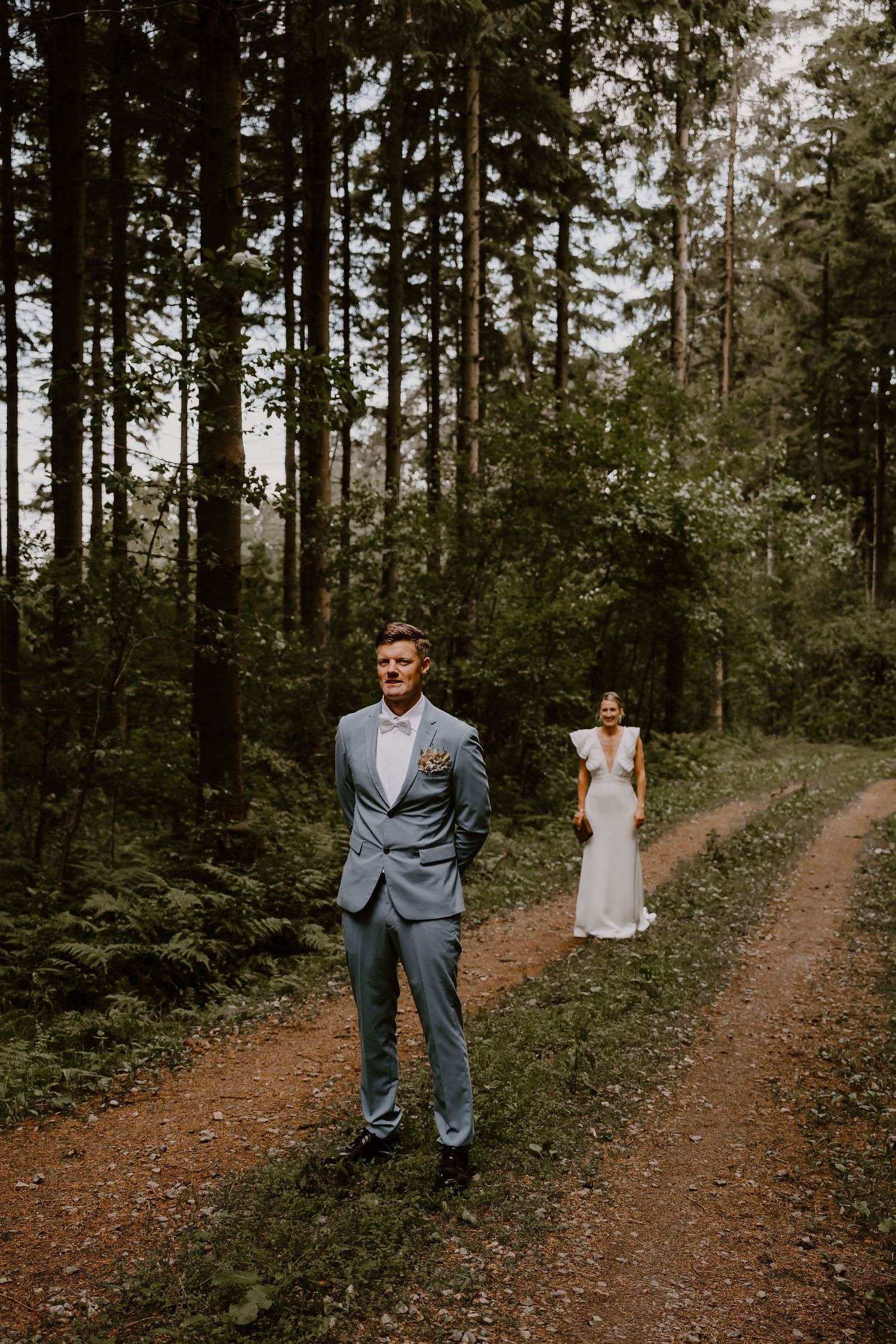 Couple posing separately on a moody forest path surrounded by tall evergreen trees during an outdoor photo session.