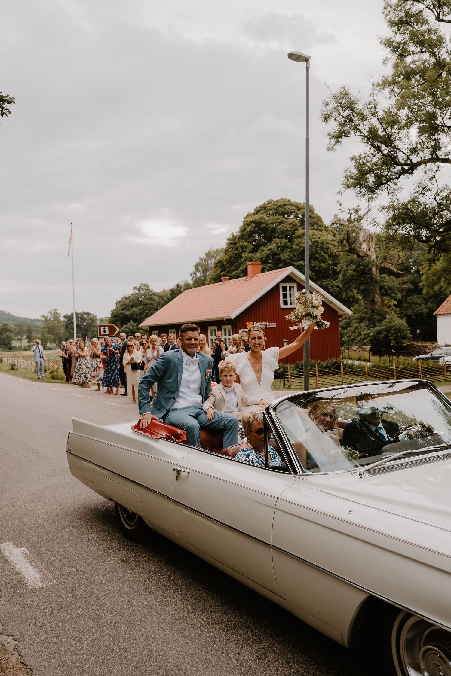 A vintage white convertible car carrying wedding guests parked along a tree-lined rural road.