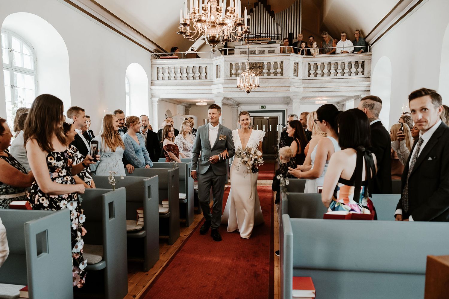 Wedding guests seated in blue church pews beneath an elegant crystal chandelier during indoor ceremony.