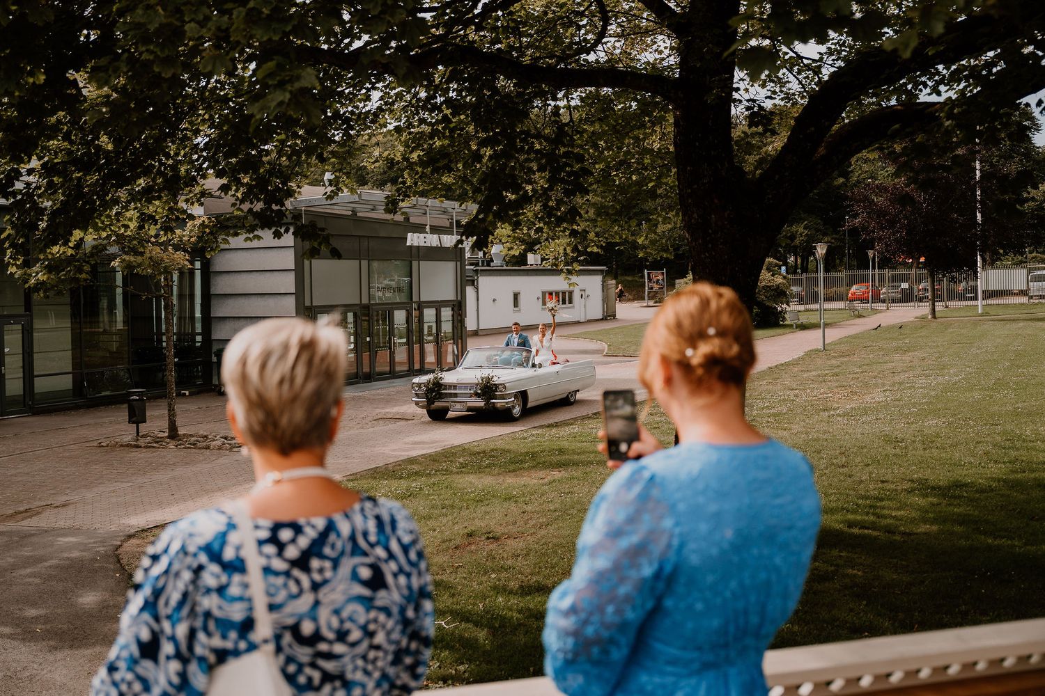 Two people in autumn attire sit watching a classic car pass by on a tree-lined street.