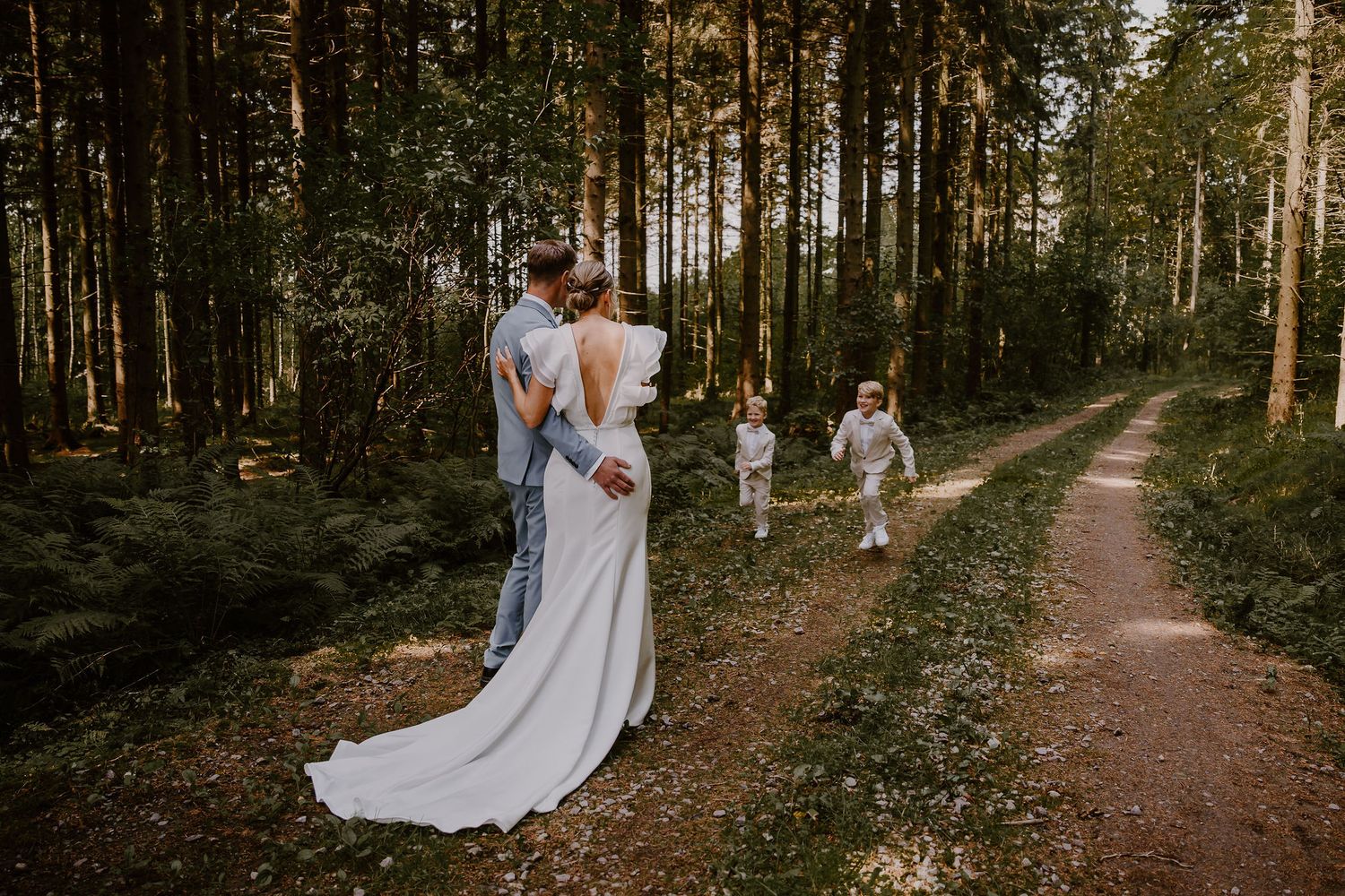 A couple embraces on a forest path while a child in white walks ahead of them down the wooded trail.